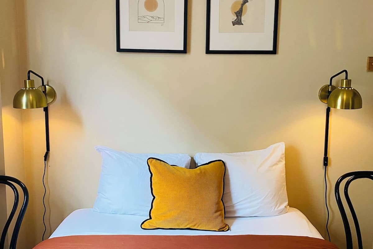 A neatly made bed with a red blanket, white pillows, and a mustard yellow accent pillow. Two framed artworks are hung on the wall above the bed.