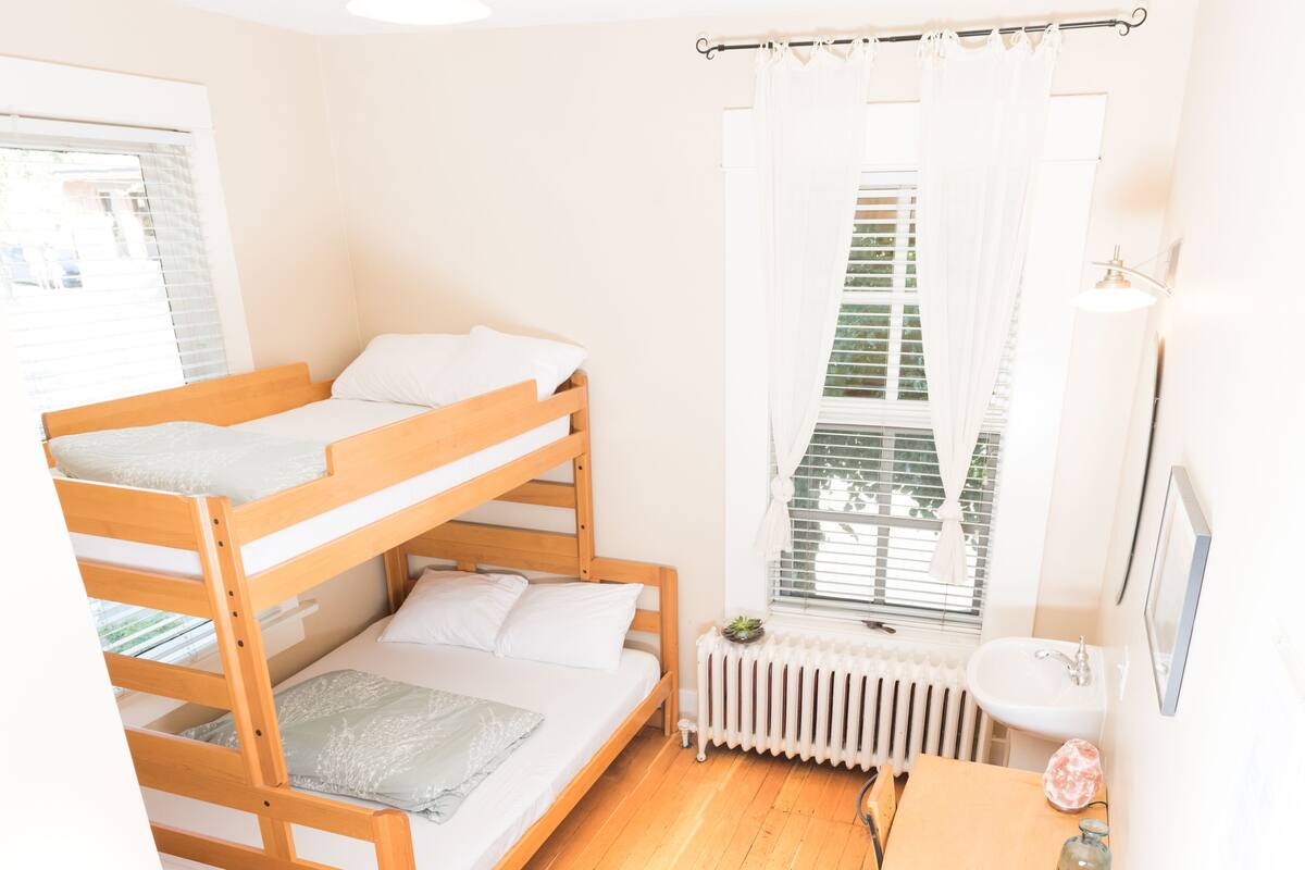 A bunk bed room with a radiator, a window with curtains, and a small table with a bottle.