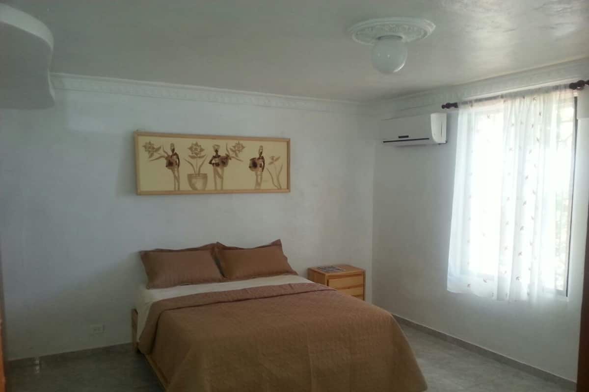 Quarto com cama, mesa de cabeceira, ar-condicionado fixado na parede e janela com cortinas.