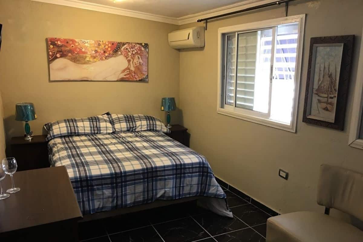 Quarto com cama, cadeira, escrivaninha com luminária, janela com persianas e obras de arte nas paredes.