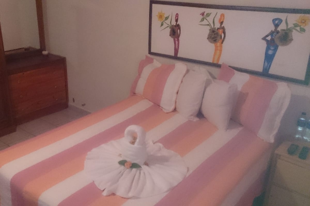 Cama bem arrumada com colcha listrada rosa e branca, toalha branca e uma boneca branca pequena. Um aparador de madeira à esquerda e uma obra de arte emoldurada acima da cama.
