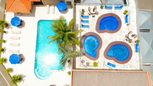 Un área de piscina en la azotea con una piscina central grande y tres jacuzzis más pequeños, rodeada de sillas de descanso y sombrillas.