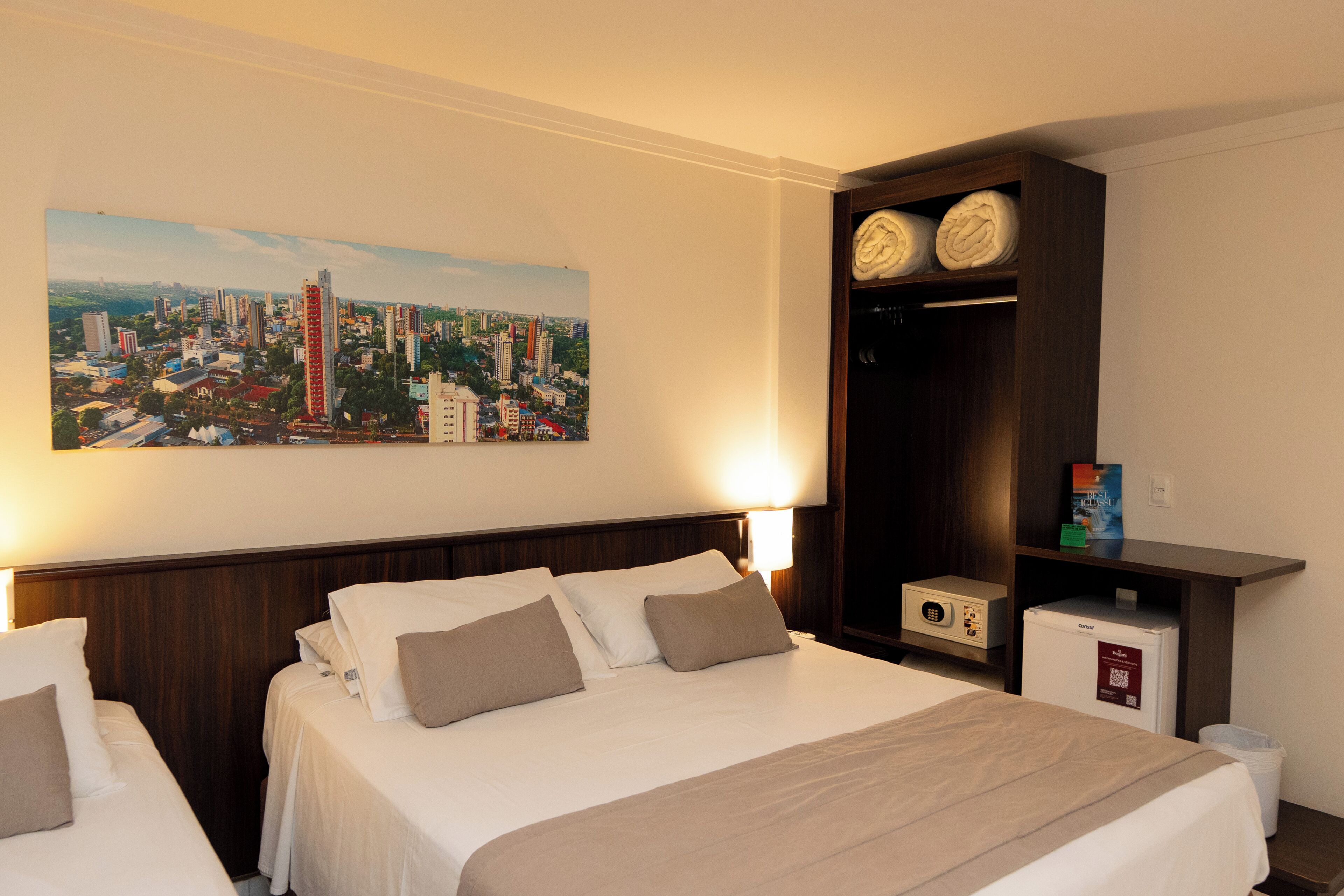 Une chambre d’hôtel comprenant un lit, une table de chevet, une lampe, une étagère avec des objets décoratifs et un tableau représentant un paysage urbain.