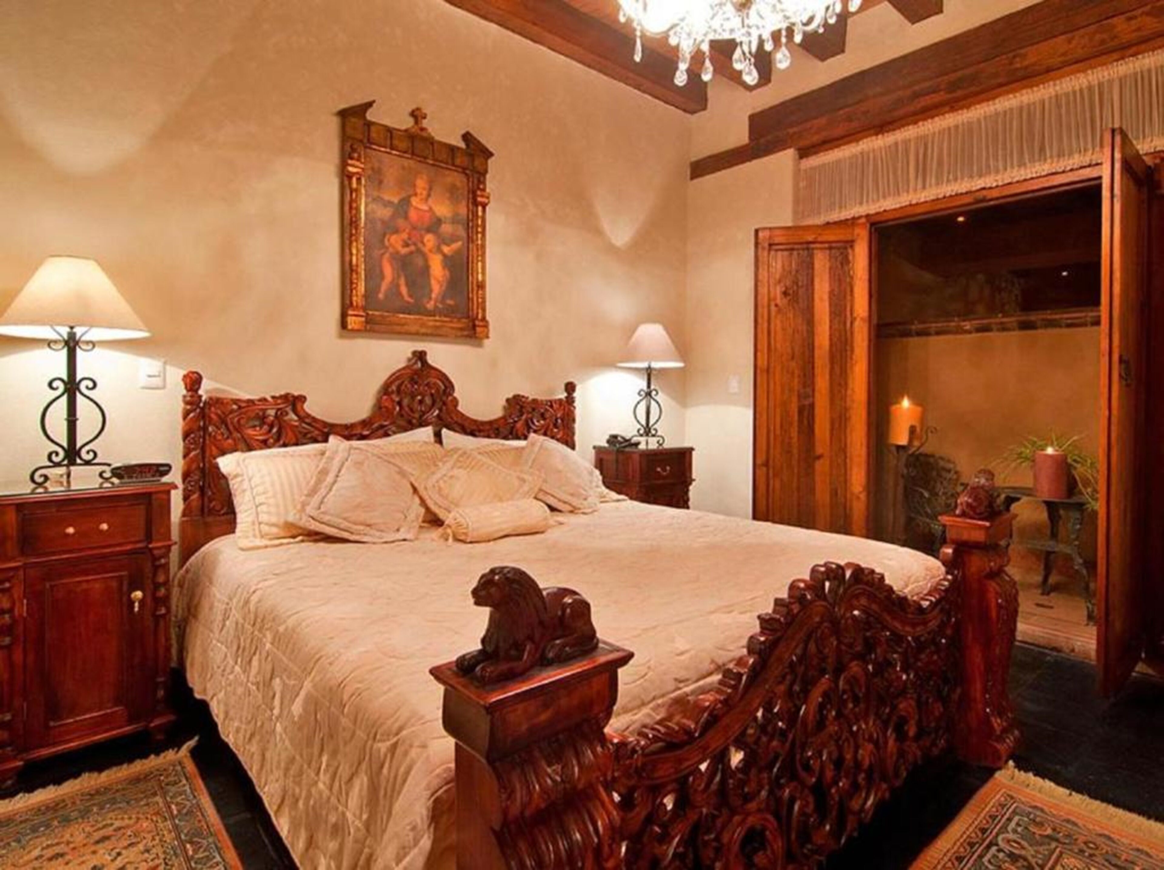Een klassieke slaapkamer met een groot bed, een gedetailleerd houten hoofdbord, twee nachtkastjes met lampen, een kroonluchter en een schilderij aan de muur.