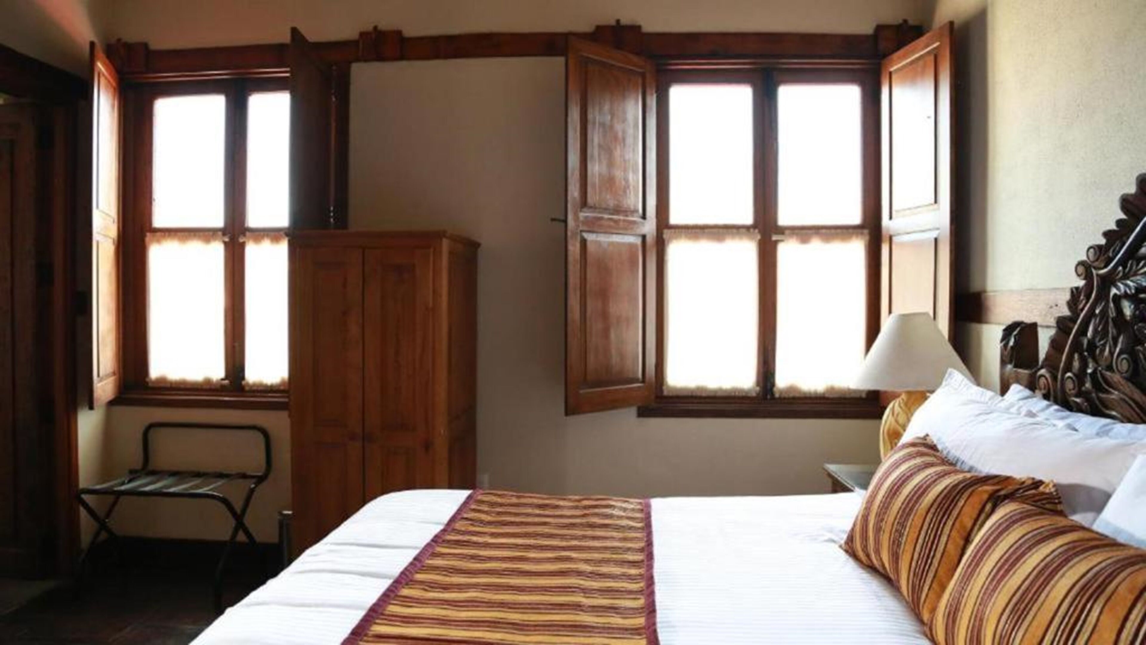 A bedroom with a bed, a nightstand, a lamp, a wardrobe, and two windows with curtains.