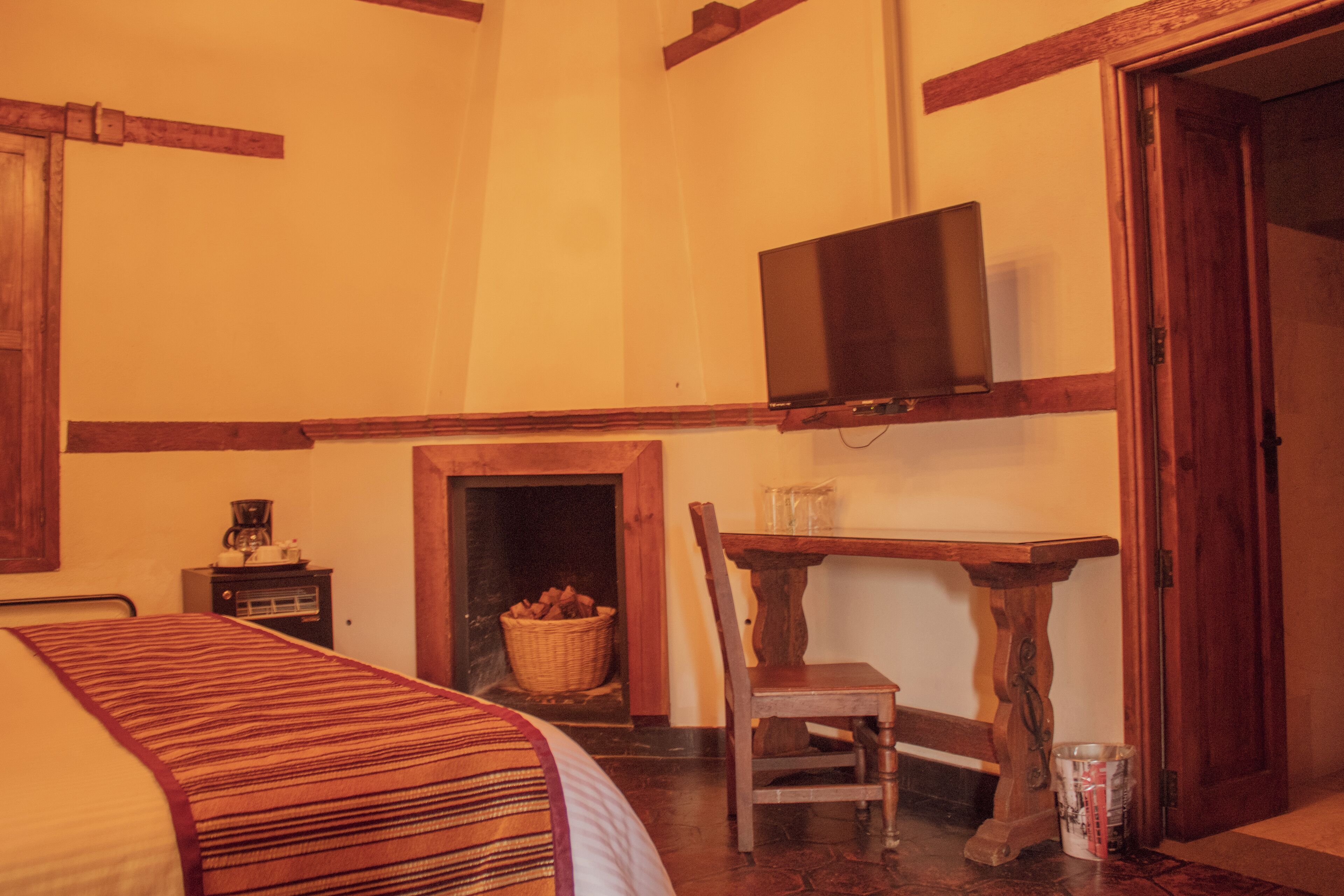 A cozy room with a bed, a television mounted on the wall, a wooden table, a chair, and a fireplace.