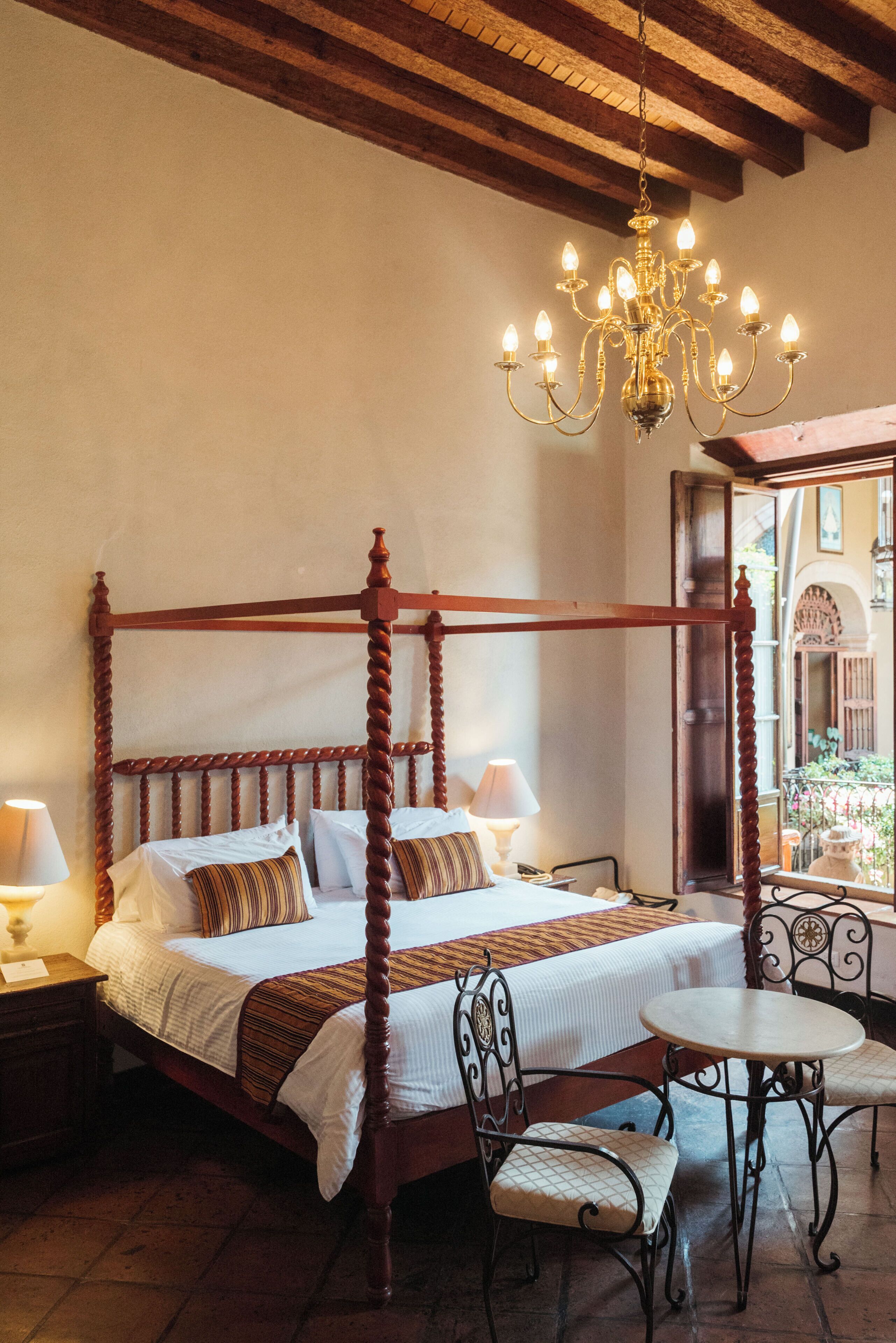 A four-poster bed with a chandelier, a small table with chairs, and a view of the courtyard through a window.