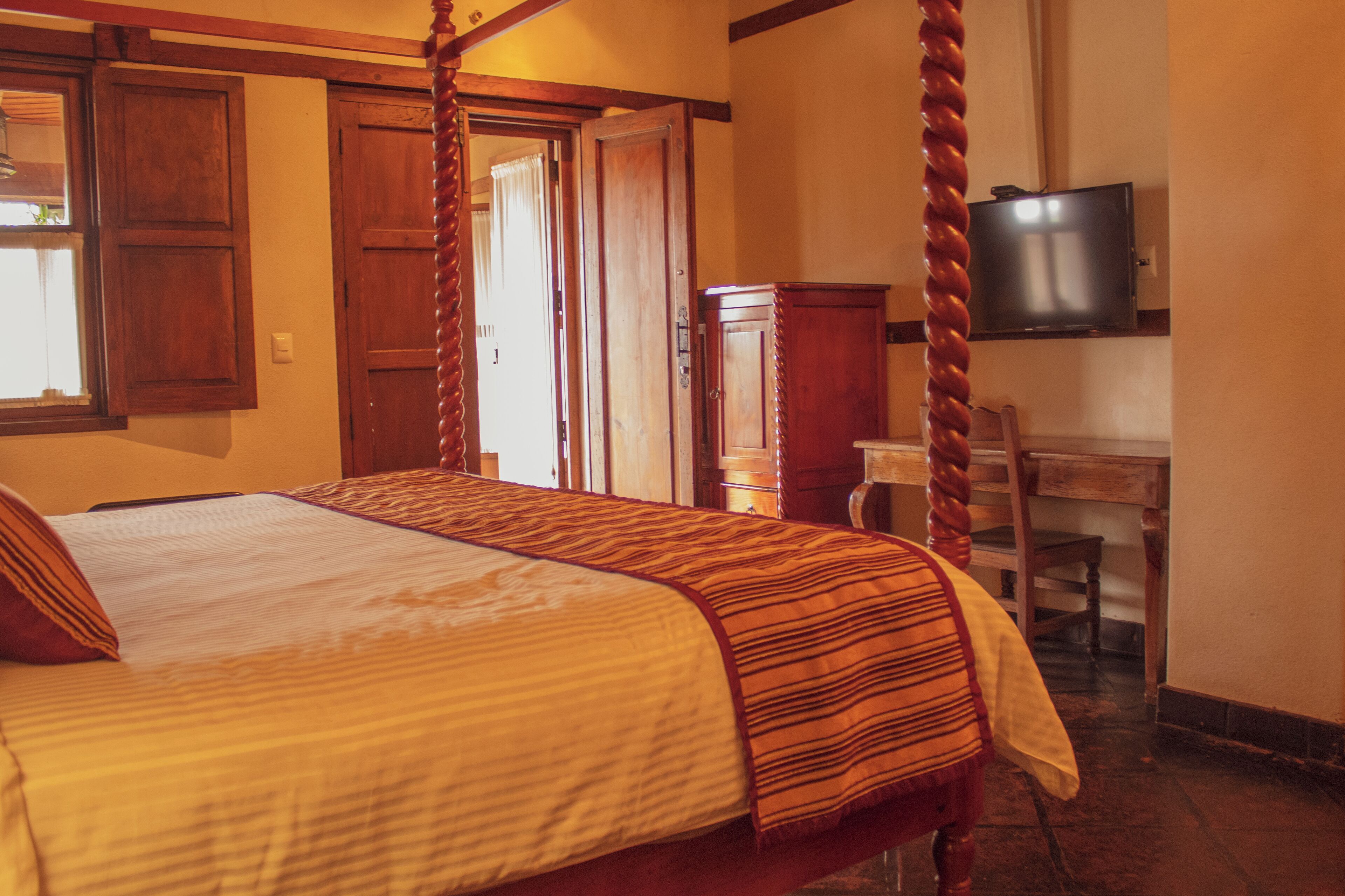 A four-poster bed with a striped bedspread, a wooden desk with a chair, and a television on the wall.