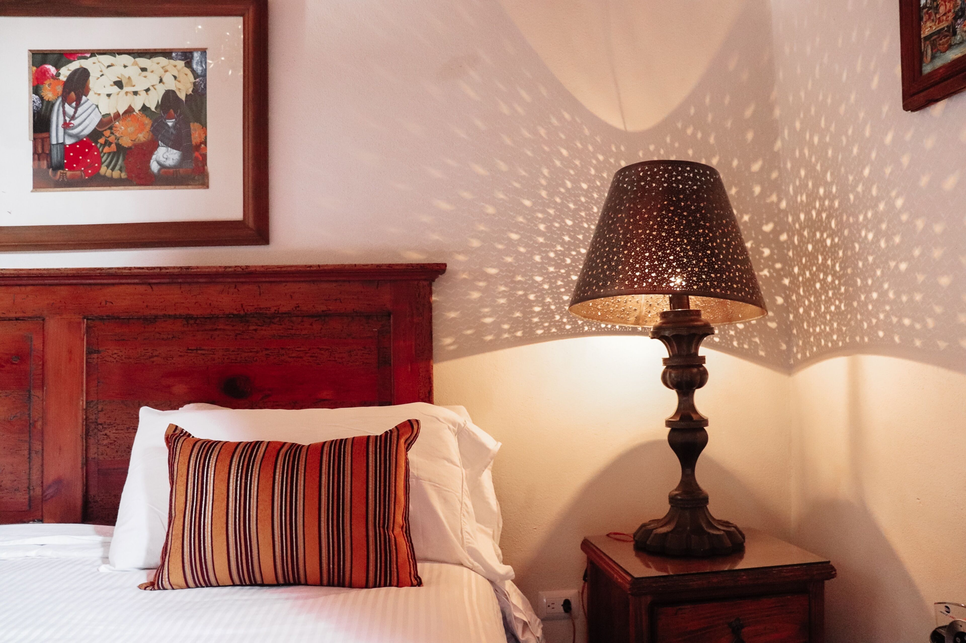 A neatly made bed with white linens and striped pillows, a wooden nightstand with a lamp, and a framed picture on the wall.