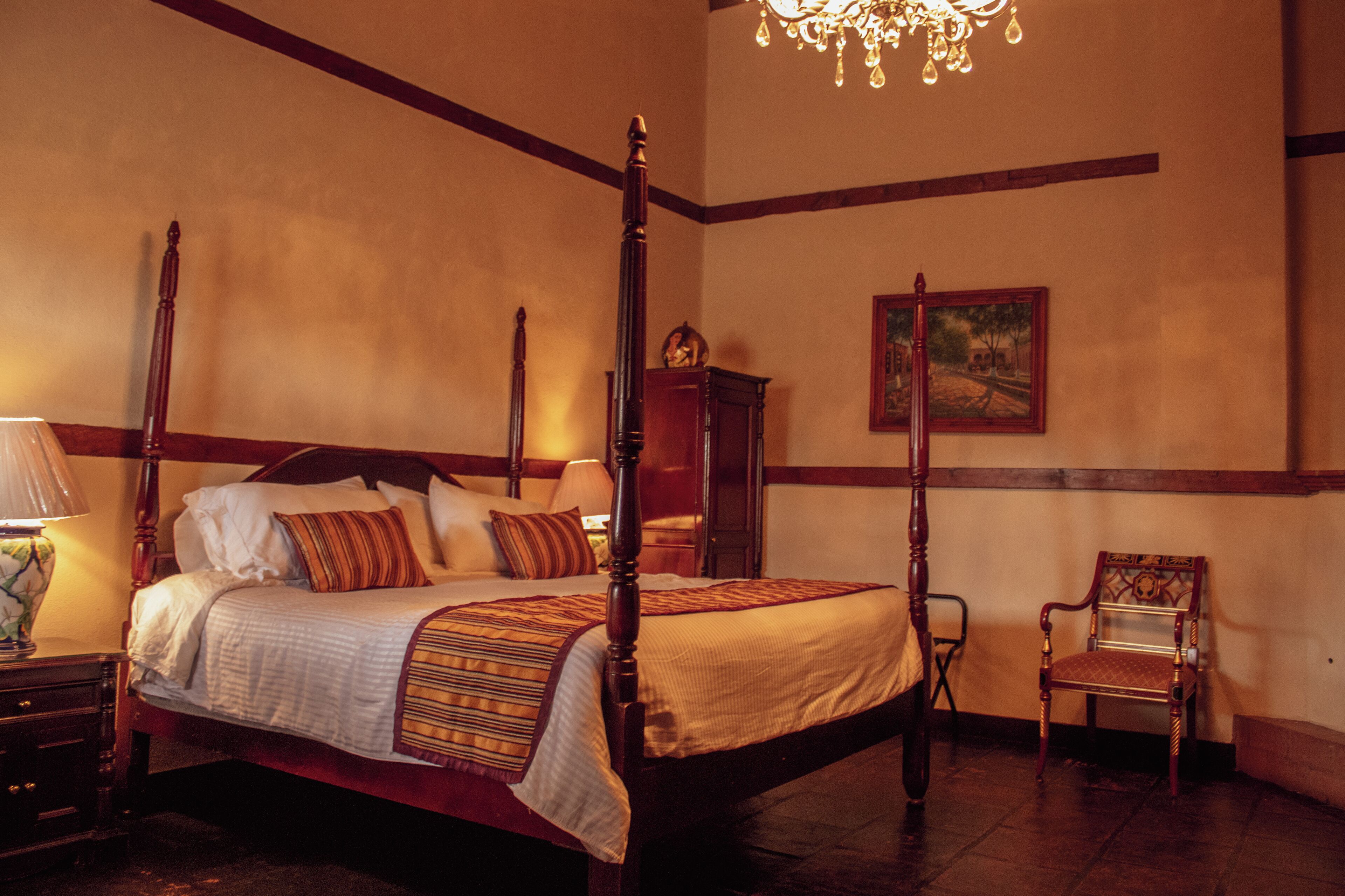 A four-poster bed with a wooden frame, a nightstand with a lamp, a chair, and a painting on the wall.