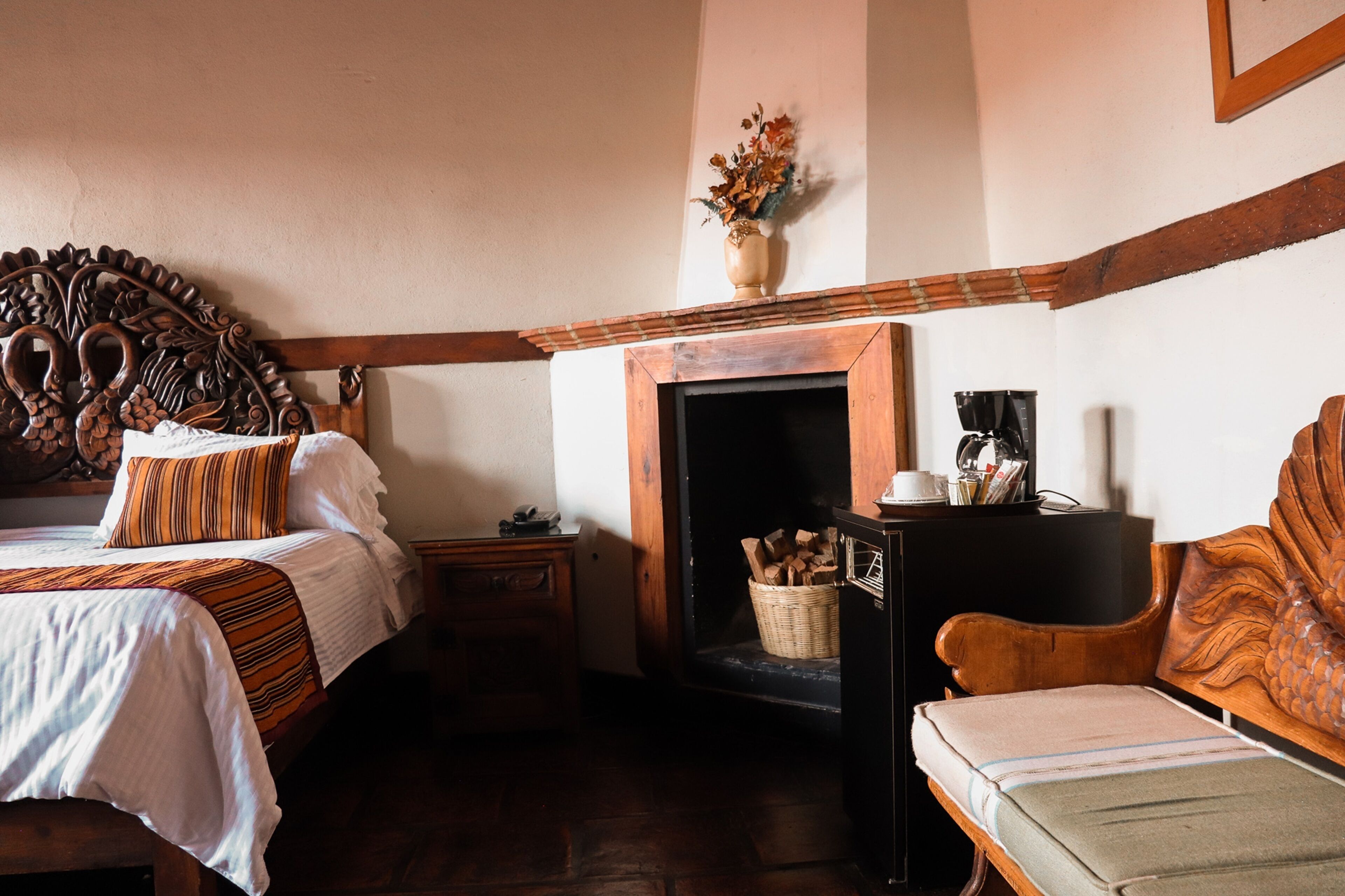 A bedroom with a bed, a nightstand, a wooden chair, a fireplace, and a vase of flowers.