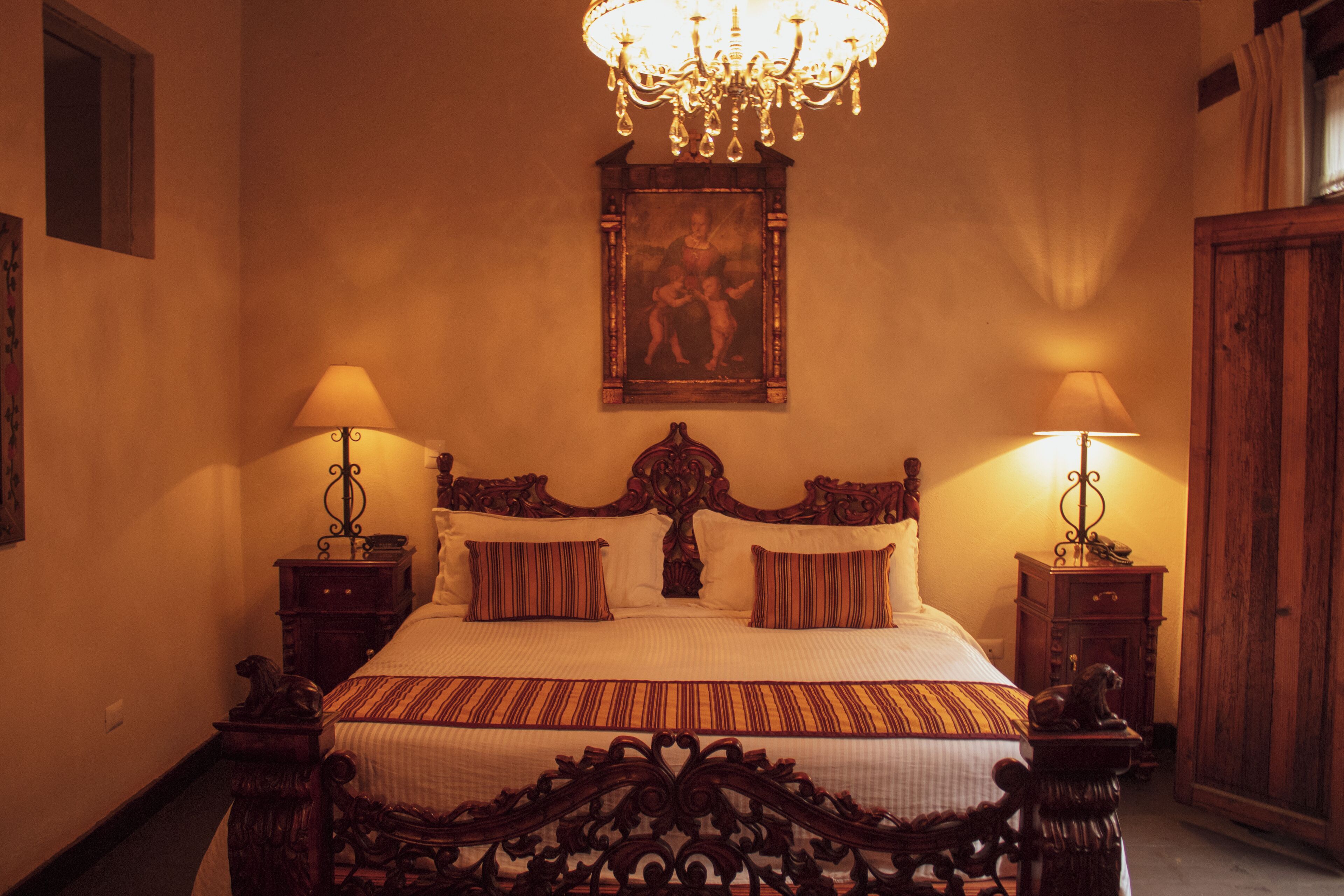 A traditional bedroom with a large bed, ornate headboard, two bedside tables with lamps, and a chandelier.