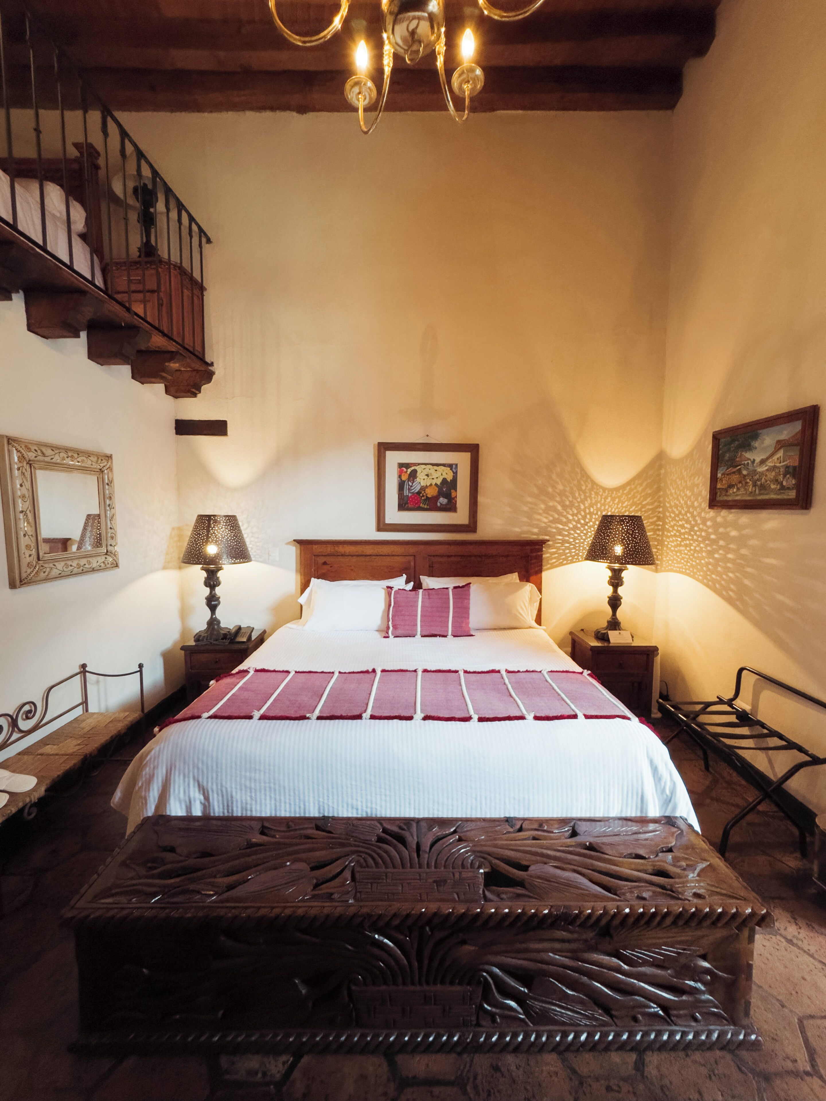 A bedroom with a large bed, two bedside tables with lamps, a wooden bench, a staircase, and two framed pictures on the walls.