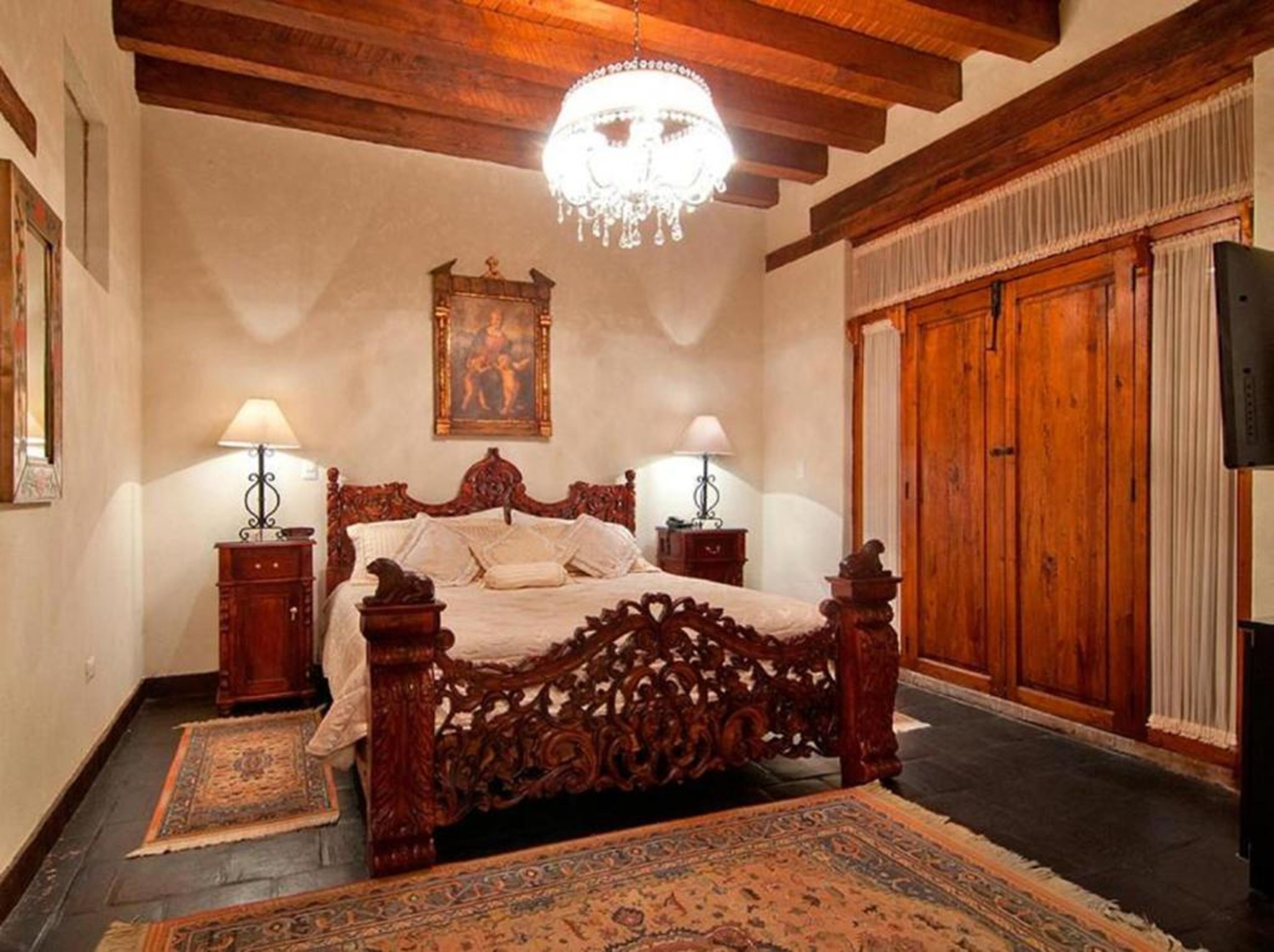 A bedroom with a wooden bed, two bedside tables, a chandelier, a painting, and a wardrobe.