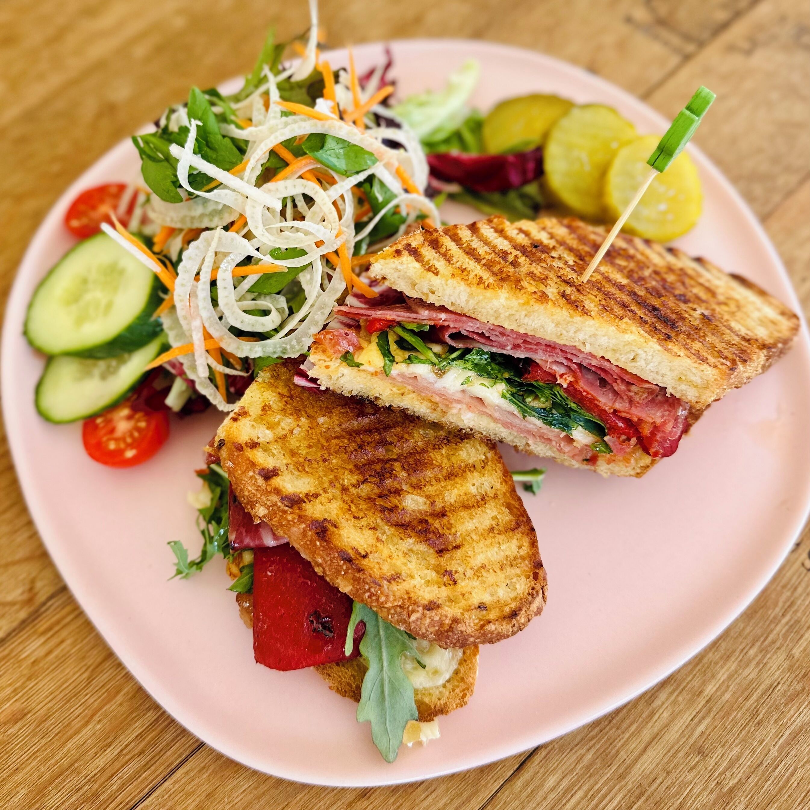 Un panino tagliato a metà su un piatto rosa, accompagnato da un'insalata e fette di cetriolo.