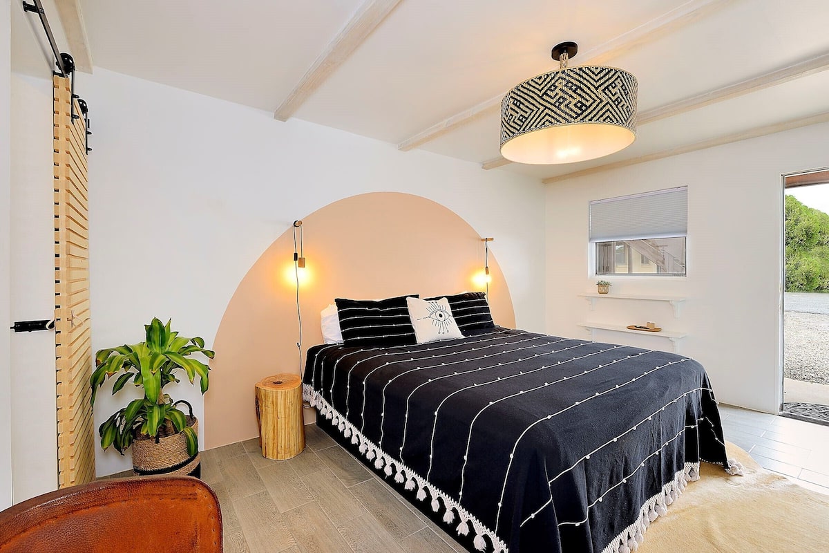 Une chambre à coucher avec un lit, des lampes de chevet, une plante en pot, un tabouret en bois et une fenêtre donnant sur un beau paysage.