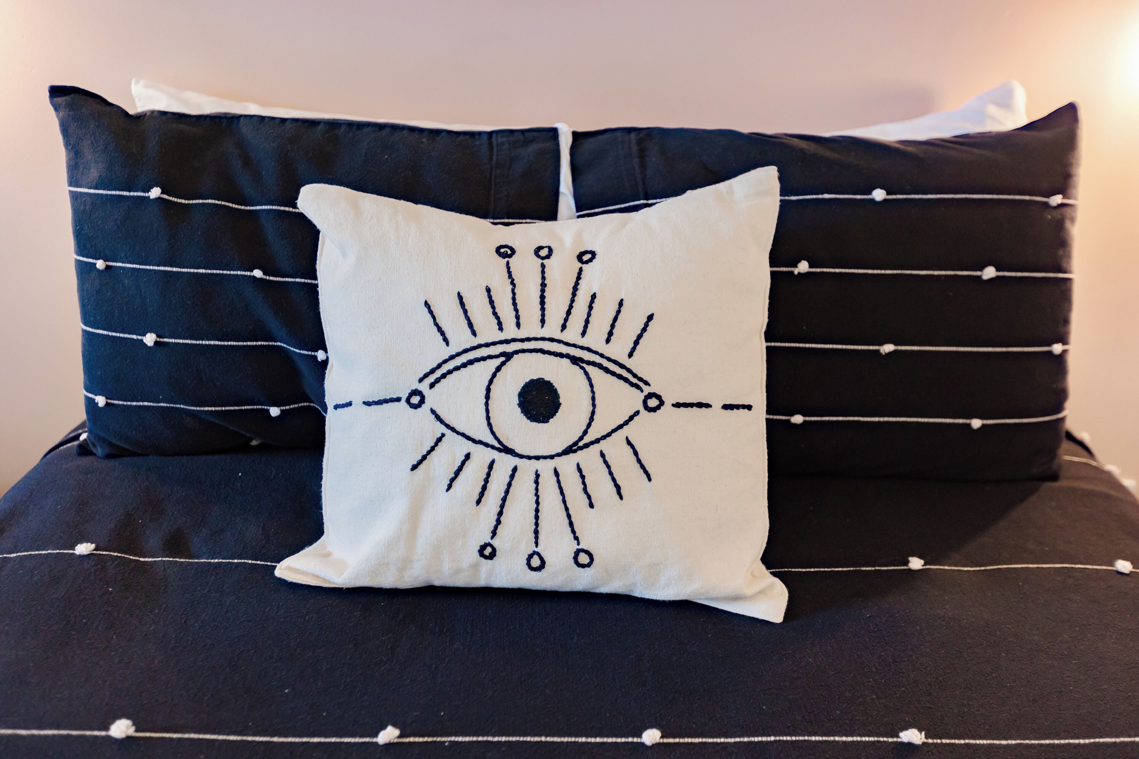 Ein Bett mit einem weißen Kopfkissen mit einem Augenmotiv, umrahmt von zwei dunklen Kissen.
