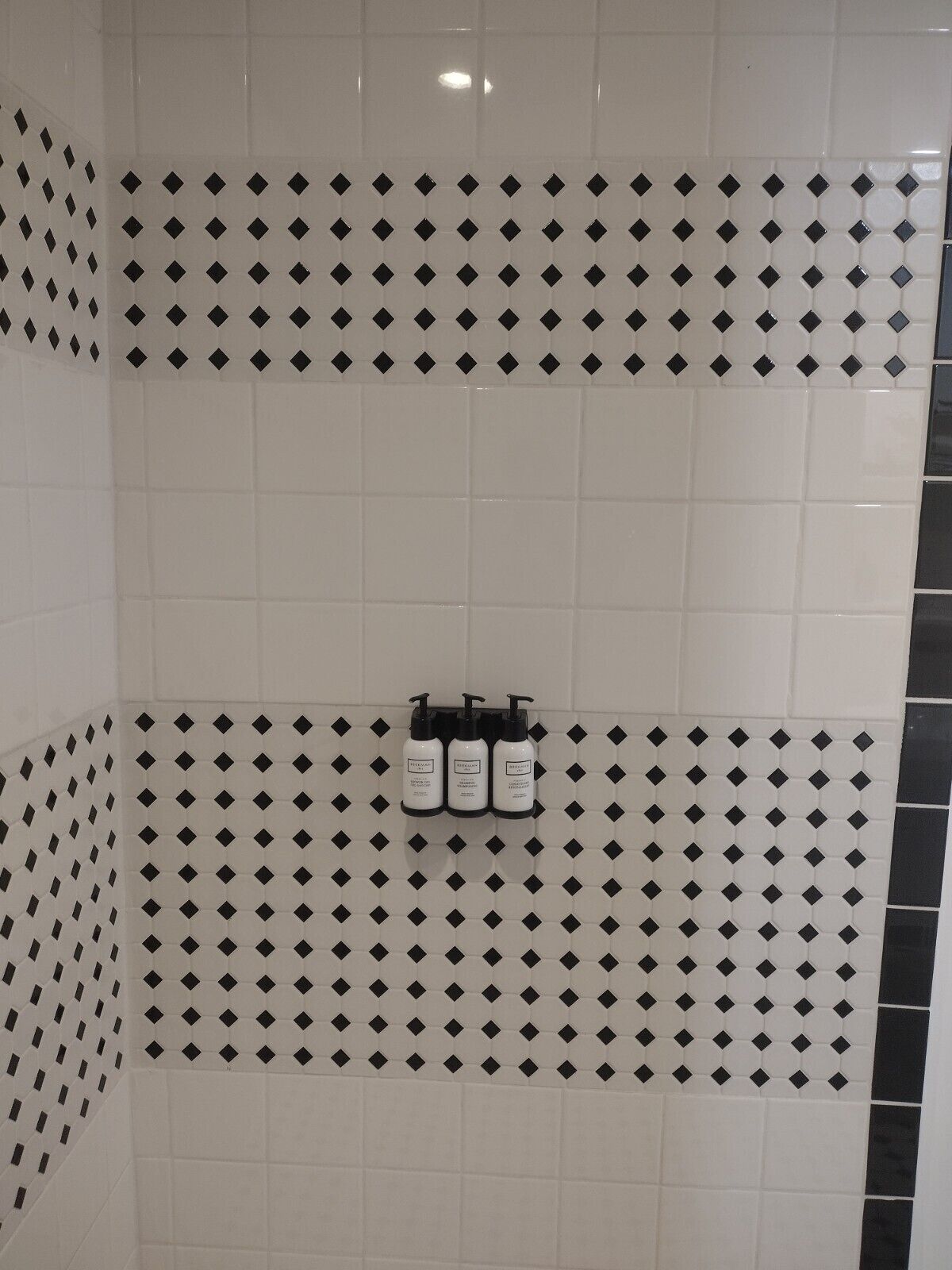A bathroom with white and black diamond-patterned tiles, a towel rack with three bottles, and a corner mirror.