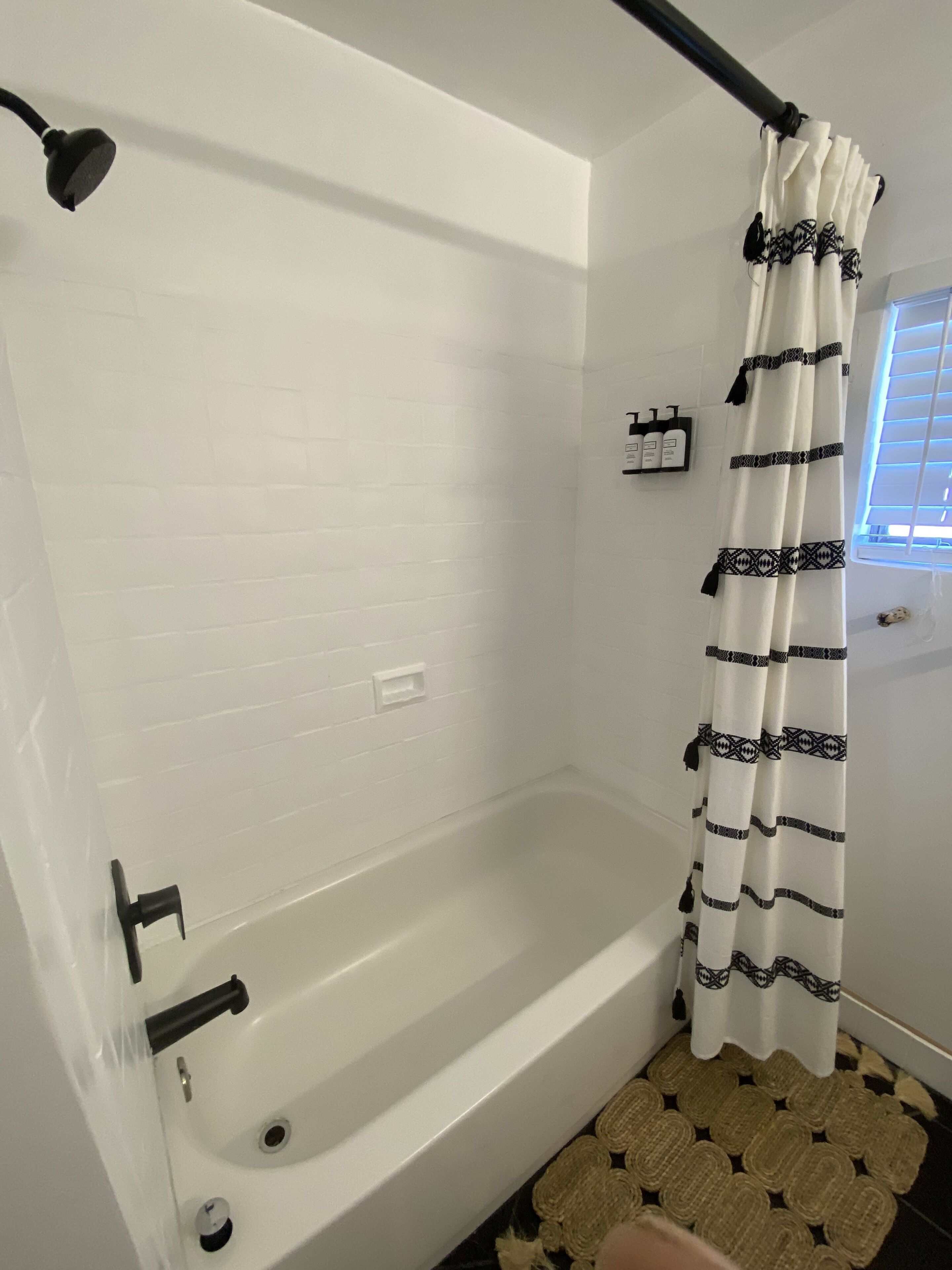 A bathroom with a white bathtub, a black showerhead, a white tiled wall, a black and white striped shower curtain, and a small shelf with toiletries.