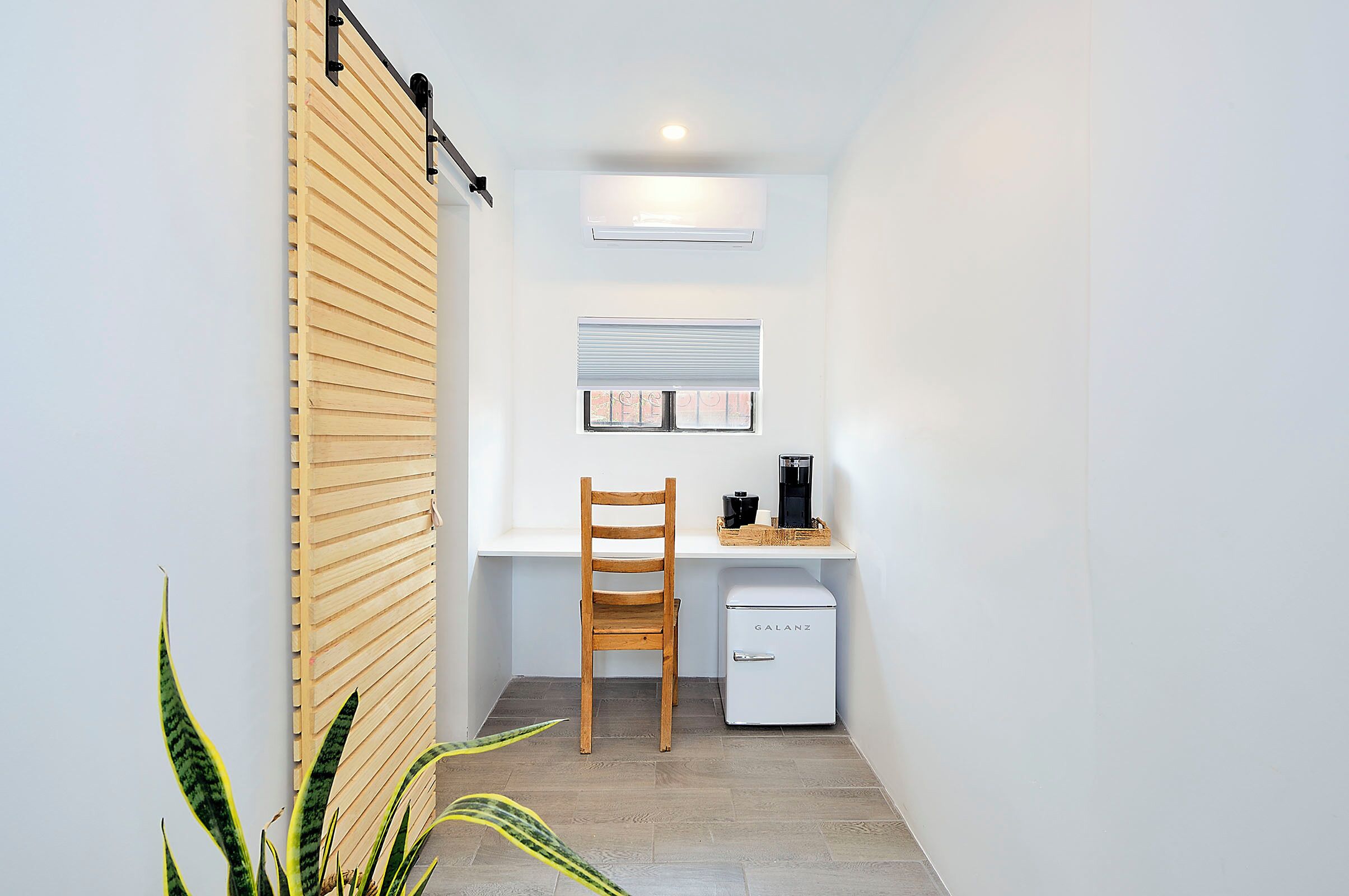 A compact kitchen with a wooden slatted wall, a white refrigerator, a wooden chair, and a small table with a coffee maker.