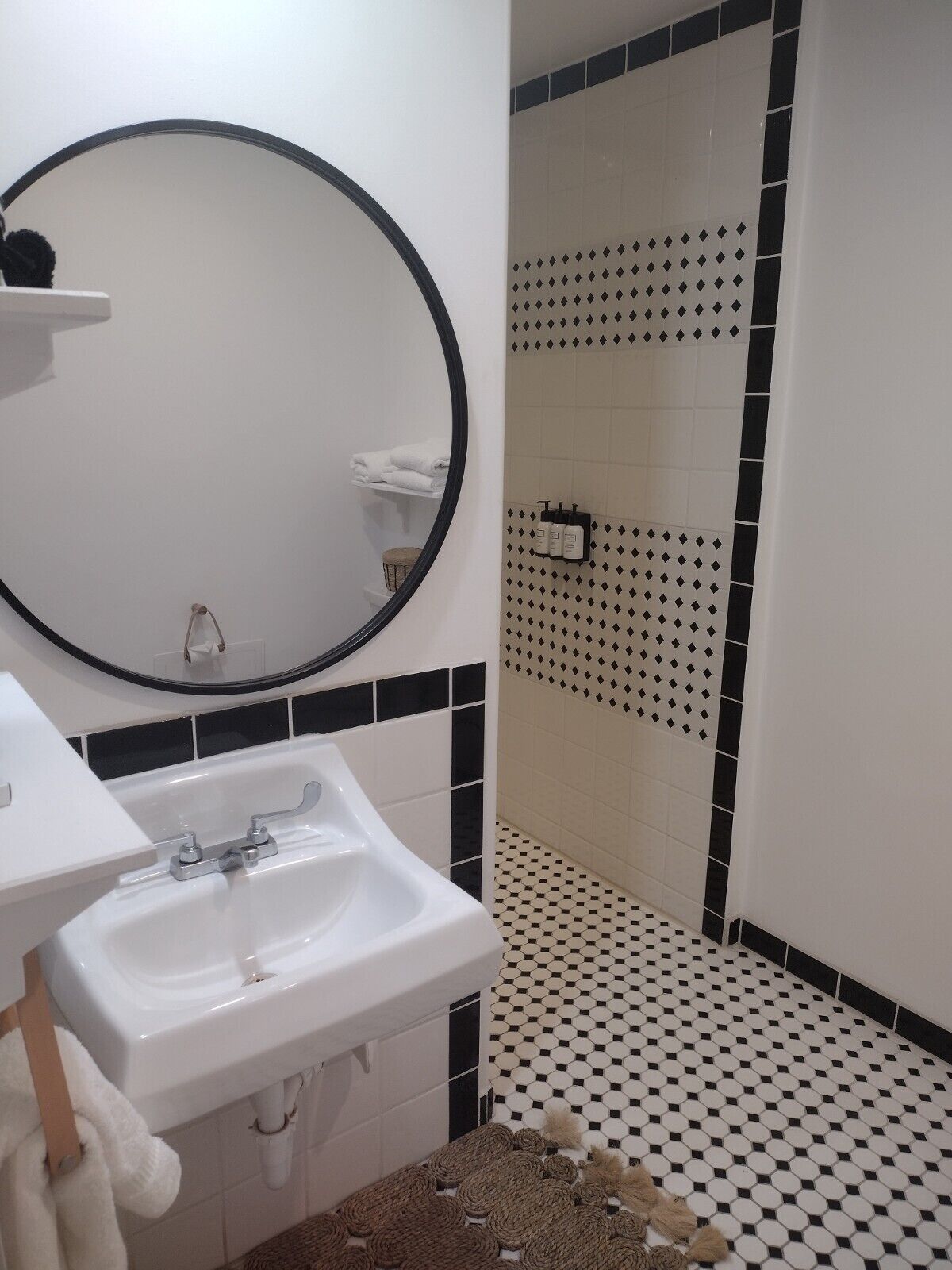 A bathroom with a white sink, a round mirror, and a shower area with black and white patterned tiles.