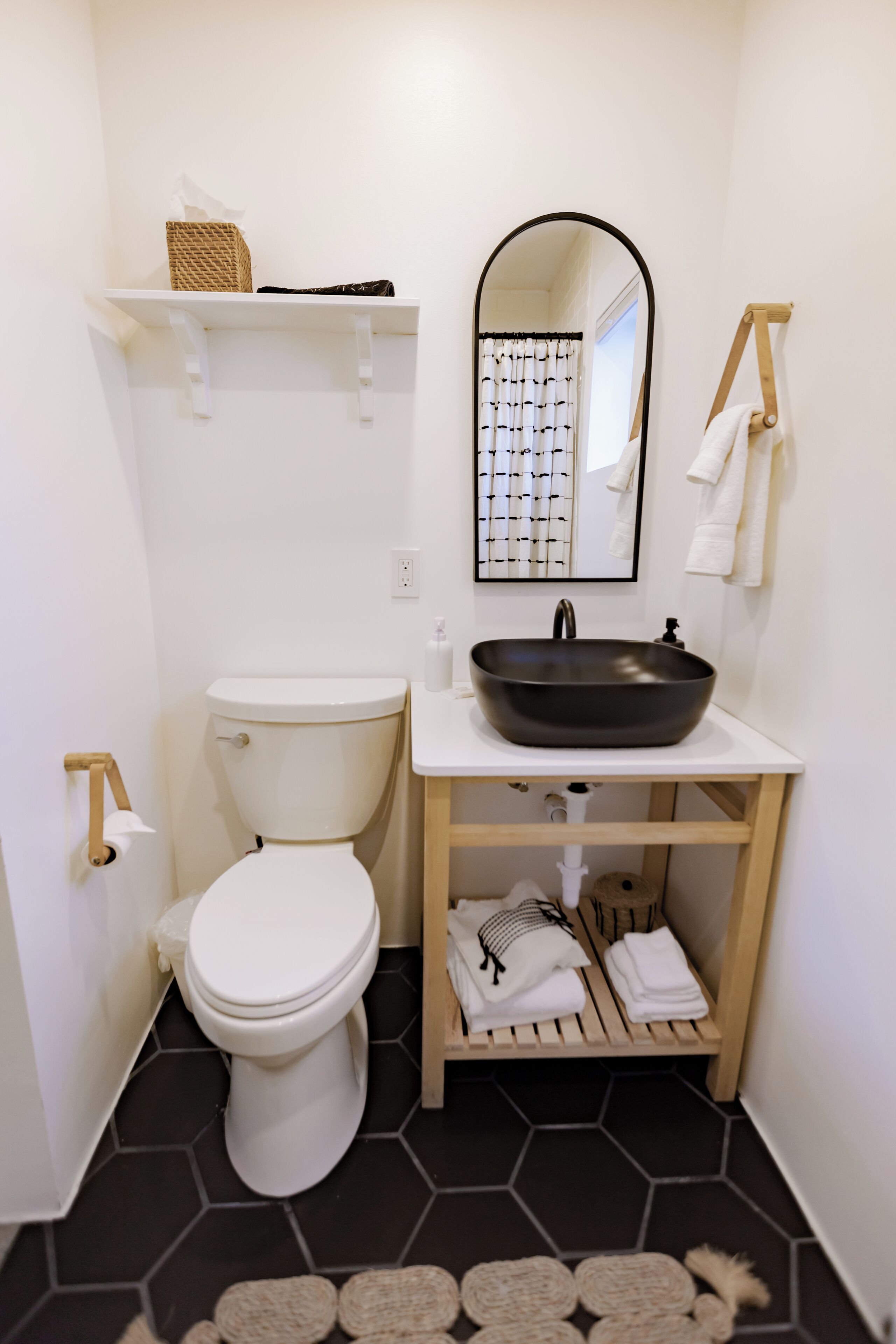 Une petite salle de bain avec des toilettes blanches, un lavabo noir et un miroir au-dessus. On y trouve des étagères avec des serviettes et un petit panier.