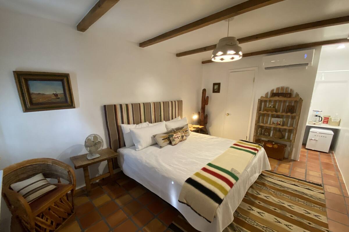Une chambre avec un grand lit, une tête de lit en bois, une table de chevet avec un ventilateur, un fauteuil à bascule en bois, un tapis à motifs et une petite kitchenette équipée d’un réfrigérateur.