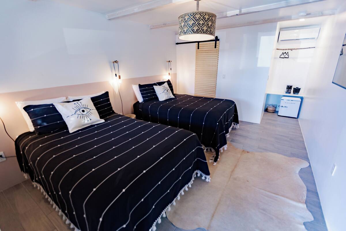 Une chambre avec deux lits, un luminaire suspendu, un petit coin cuisine et un tapis en peau de vache.
