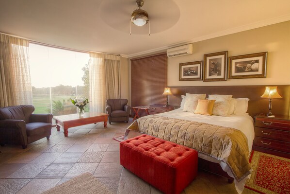 A hotel room with a large bed, a red ottoman, a chair, a small table, and a view of the outdoors.