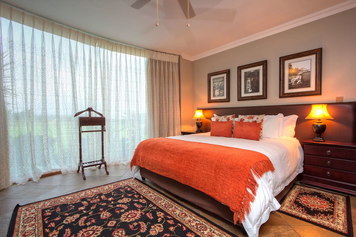 A bedroom with a large bed, two bedside lamps, a wooden nightstand, a hanging rack, and a patterned rug.
