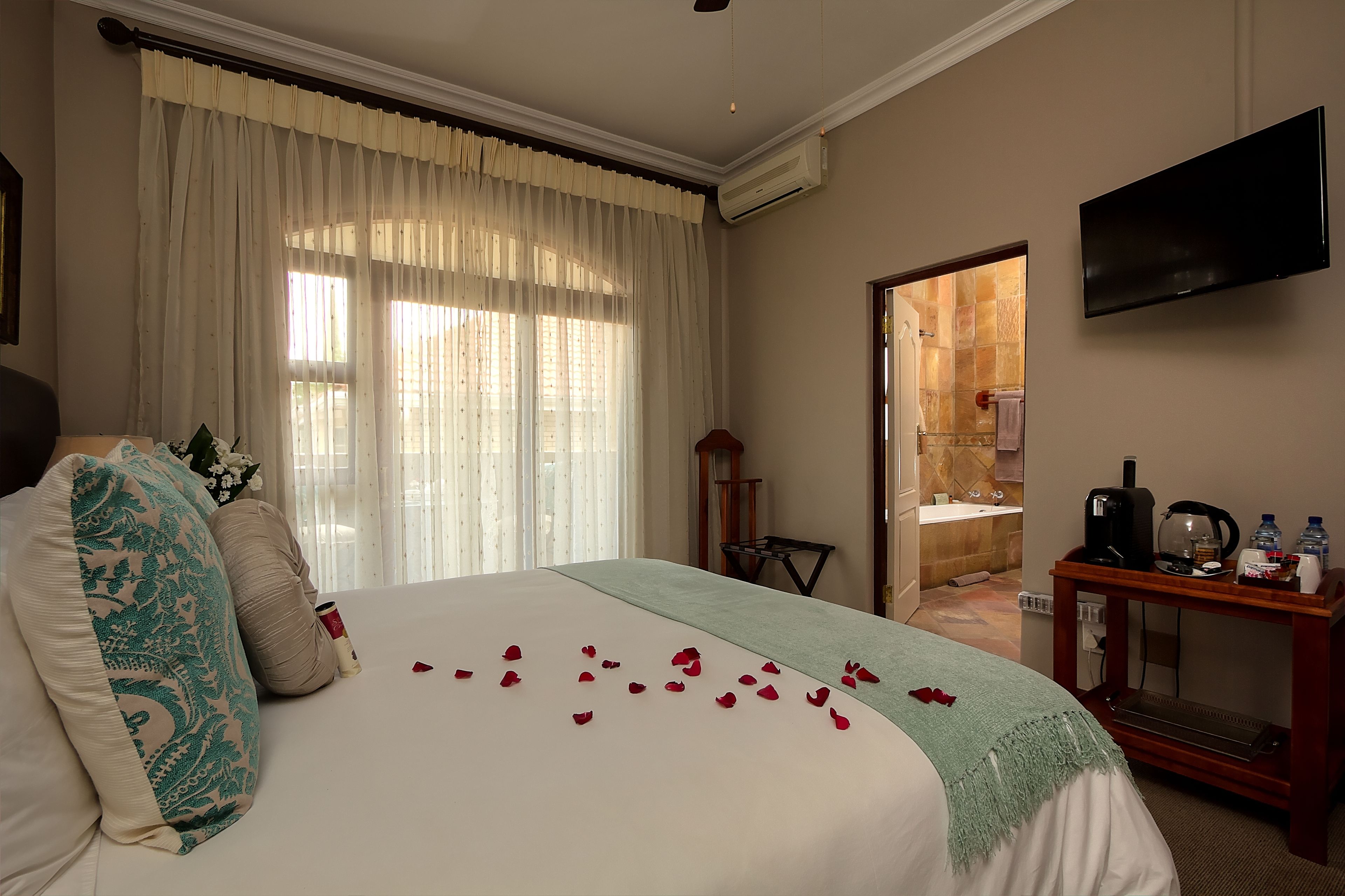 A hotel room with a bed, a bedside table with a kettle and a bottle, a TV mounted on the wall, and a bathroom visible through an open door.