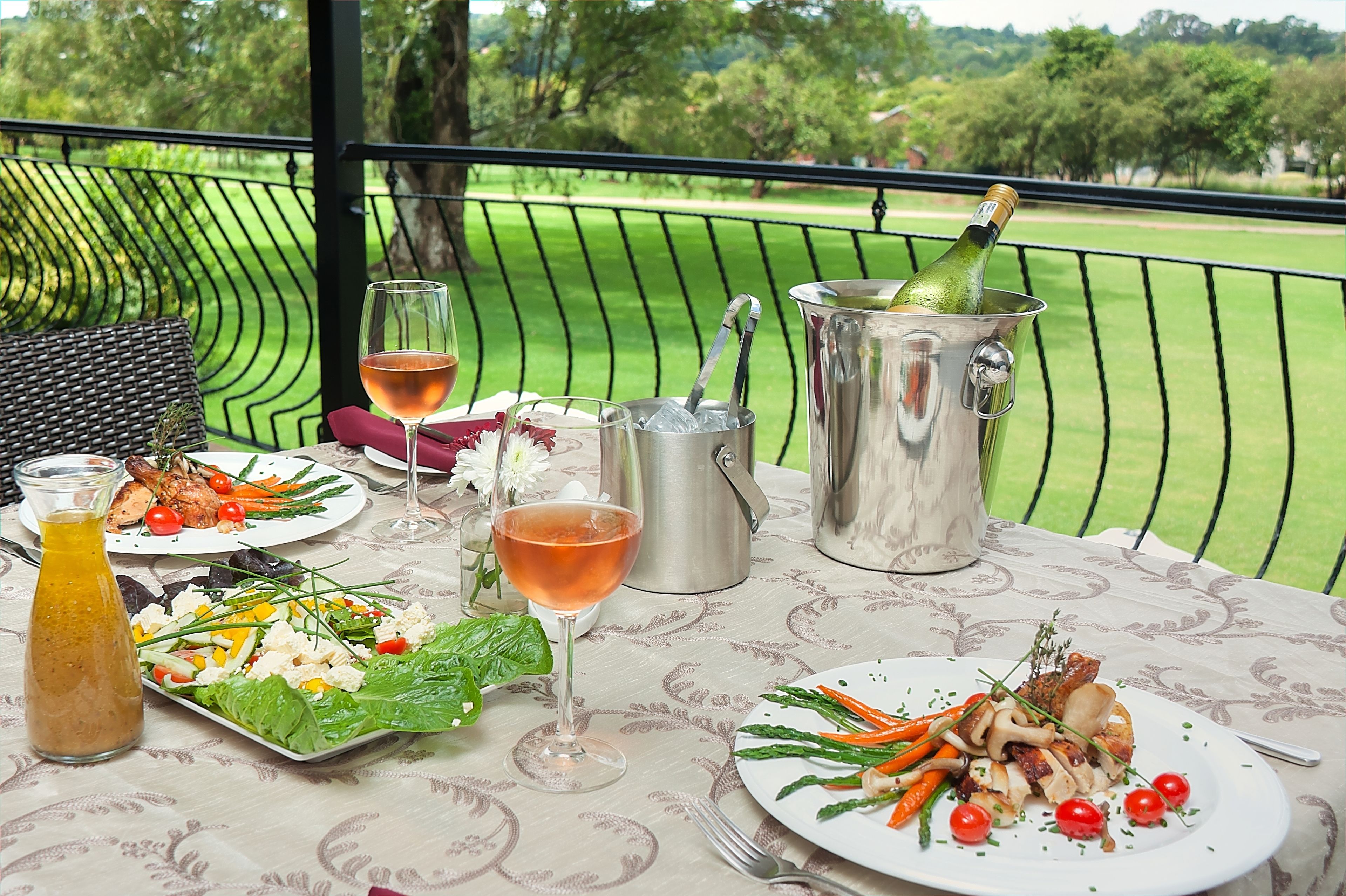 Una mesa servida con diversos platos, bebidas y una botella de champán en un balcón con vista a un campo de golf.