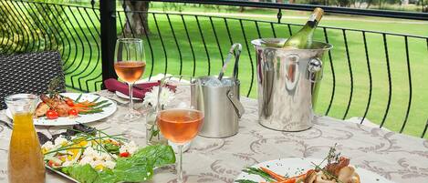 Een gedekte tafel met diverse gerechten, drankjes en een fles champagne op een balkon met uitzicht over een golfbaan.
