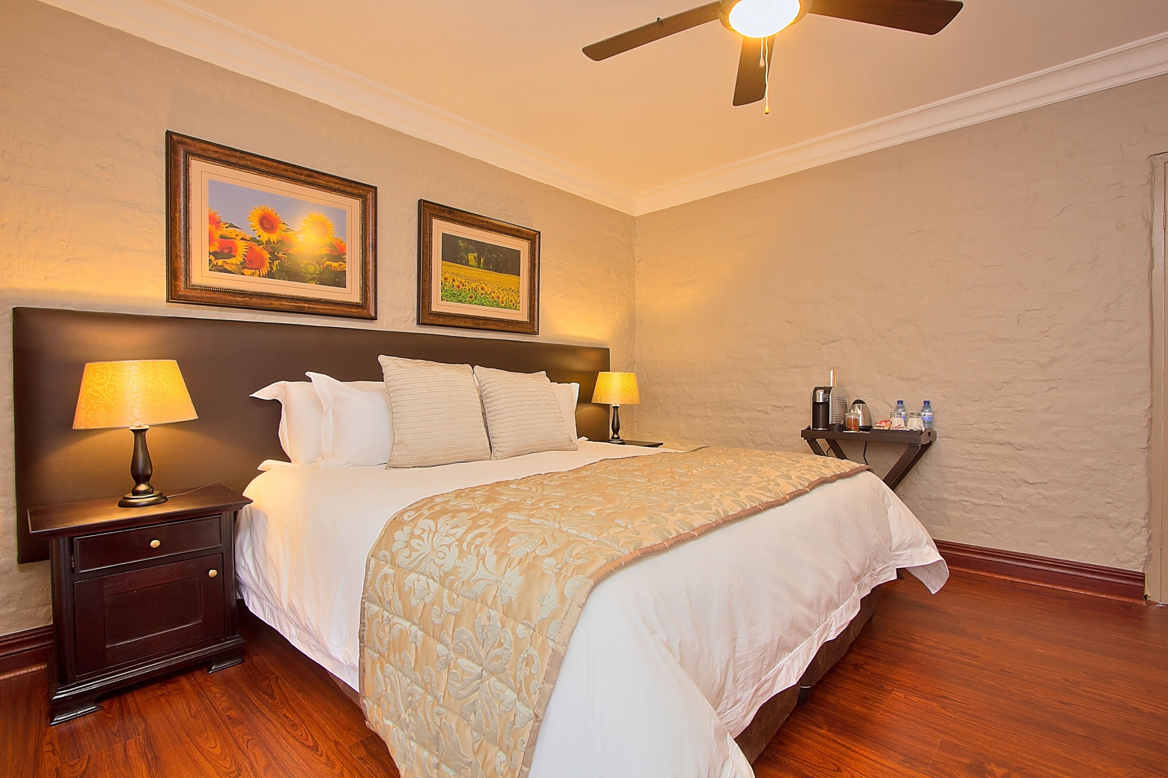 A neatly made bed with a beige bedspread, two bedside lamps, a nightstand with a lamp, and a ceiling fan.