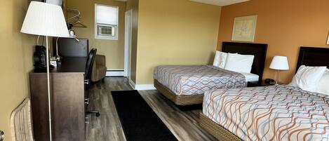 A hotel room with two beds, a desk with a lamp, a radiator, and a black rug.