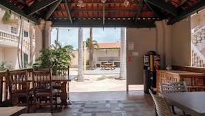 Un patio al aire libre con mesas y sillas de madera, suelo de baldosas y vista a un edificio con tejado de tejas rojas al fondo.