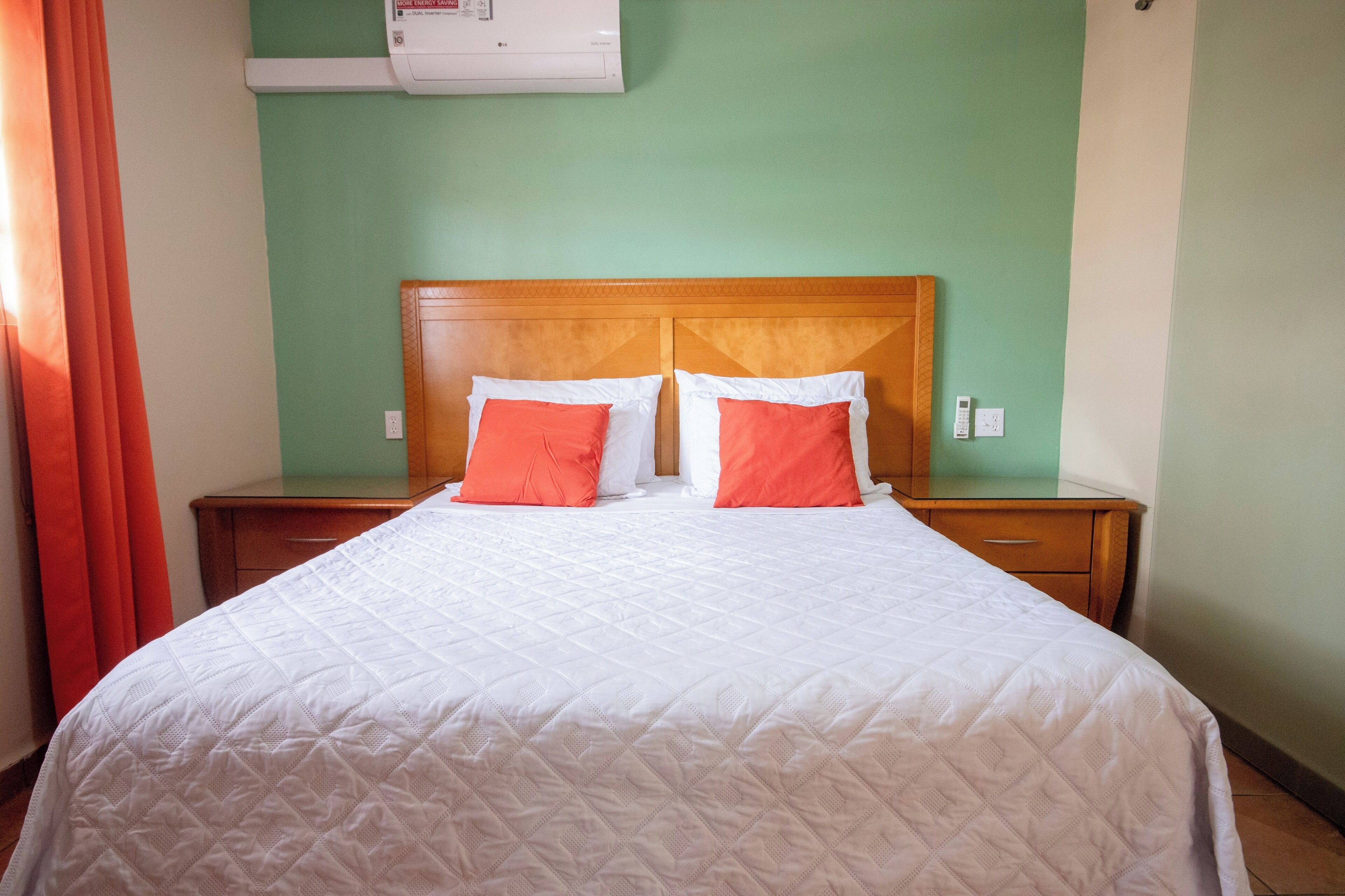 A neatly made bed with white bedding and two orange pillows, a wooden headboard, and a green wall.