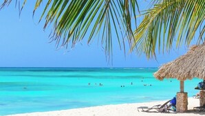 Una playa con agua turquesa cristalina, una sombrilla de paja y algunas personas a lo lejos.