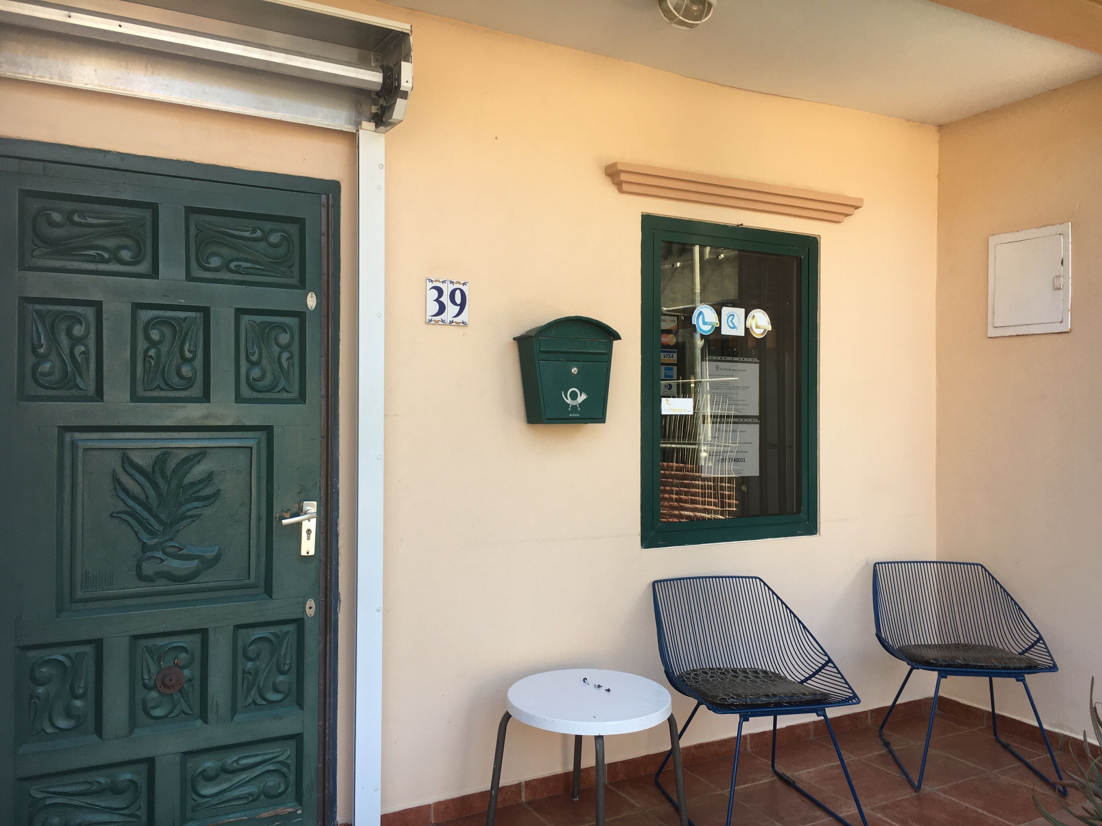Eine grüne Tür mit dekorativen Feldern, ein Briefkasten, der Hinweisschild Nummer 39 und zwei blaue Stühle mit einem kleinen weißen Tisch.