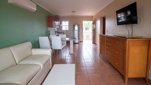 Un salón amplio con sofá, una mesa de comedor con sillas, una zona de cocina con refrigerador y un televisor montado en la pared.