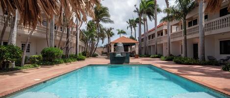 A swimming pool with a thatched roof structure, surrounded by palm trees and a multi-story building with balconies.