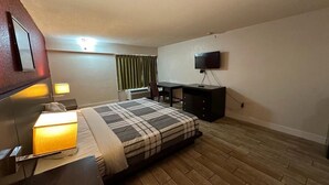 Ett hotellrum med en säng, sänglampor, ett skrivbord, en stol, en tv och ett fönster med gardiner.