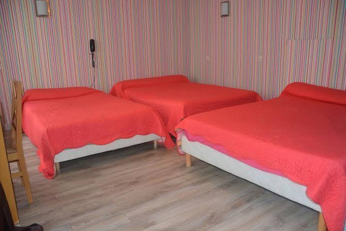 A room with three beds, each with a red blanket. The room has wooden flooring and striped wallpaper.