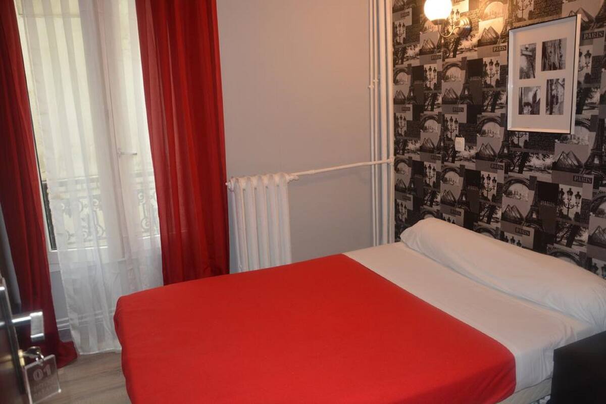 A single bed with a red bedspread in a room with a radiator, a window with red curtains, and a wall decorated with Paris-themed pictures.