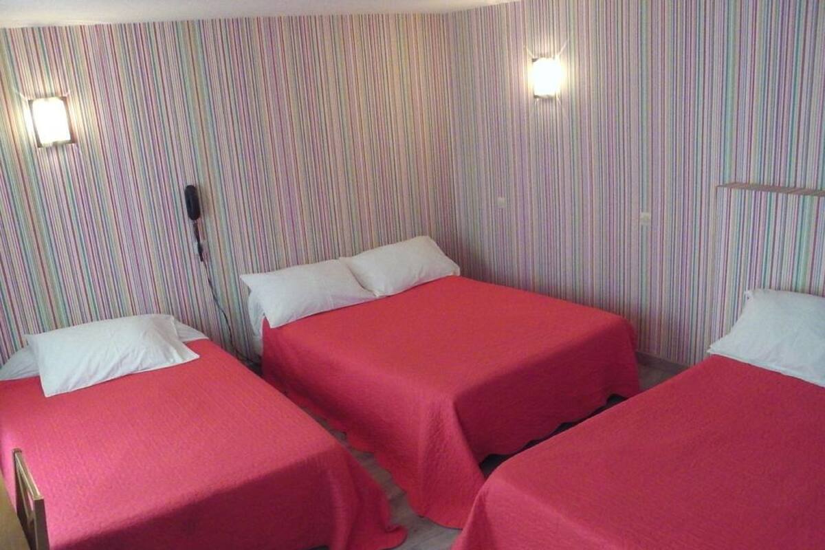 A hotel room with three single beds, each with a red bedspread and white pillows. The walls have a striped pattern, and there are two wall-mounted light fixtures.