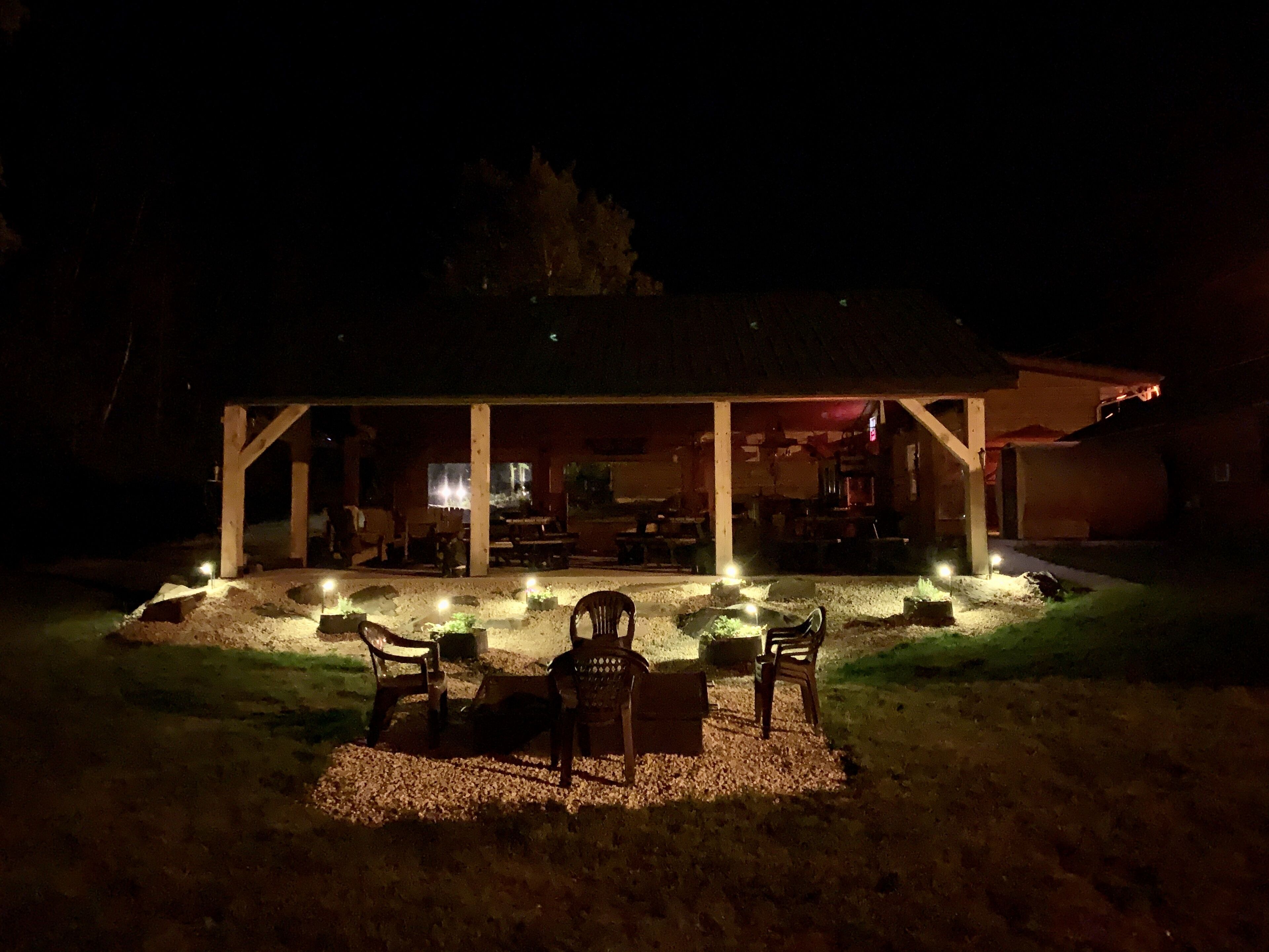 A covered outdoor seating area with chairs and tables, illuminated by ground lights, and a building in the background.
