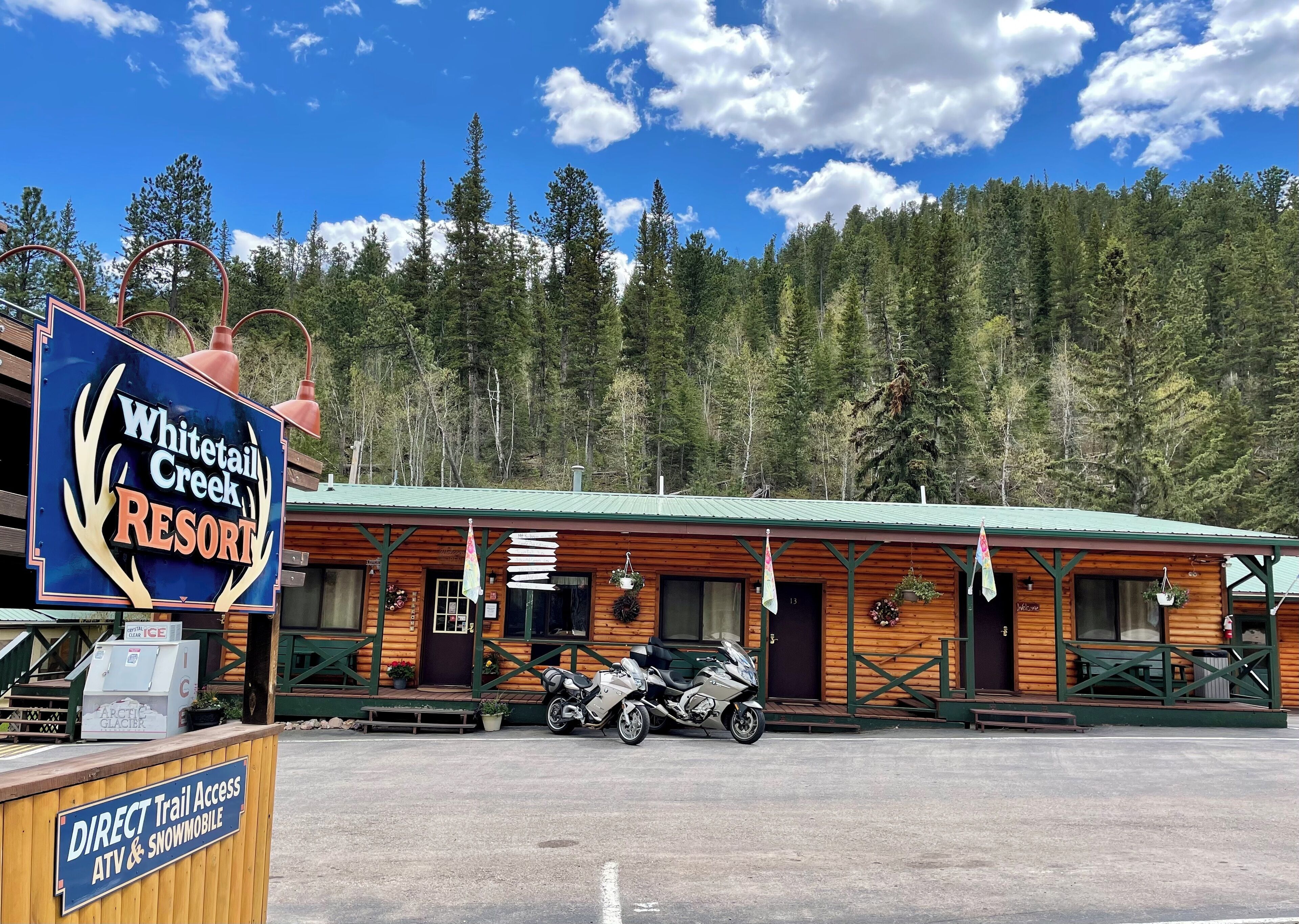 Whitetail Creek Resort with a direct trail access sign, a motorcycle parked in front, and a forested backdrop.