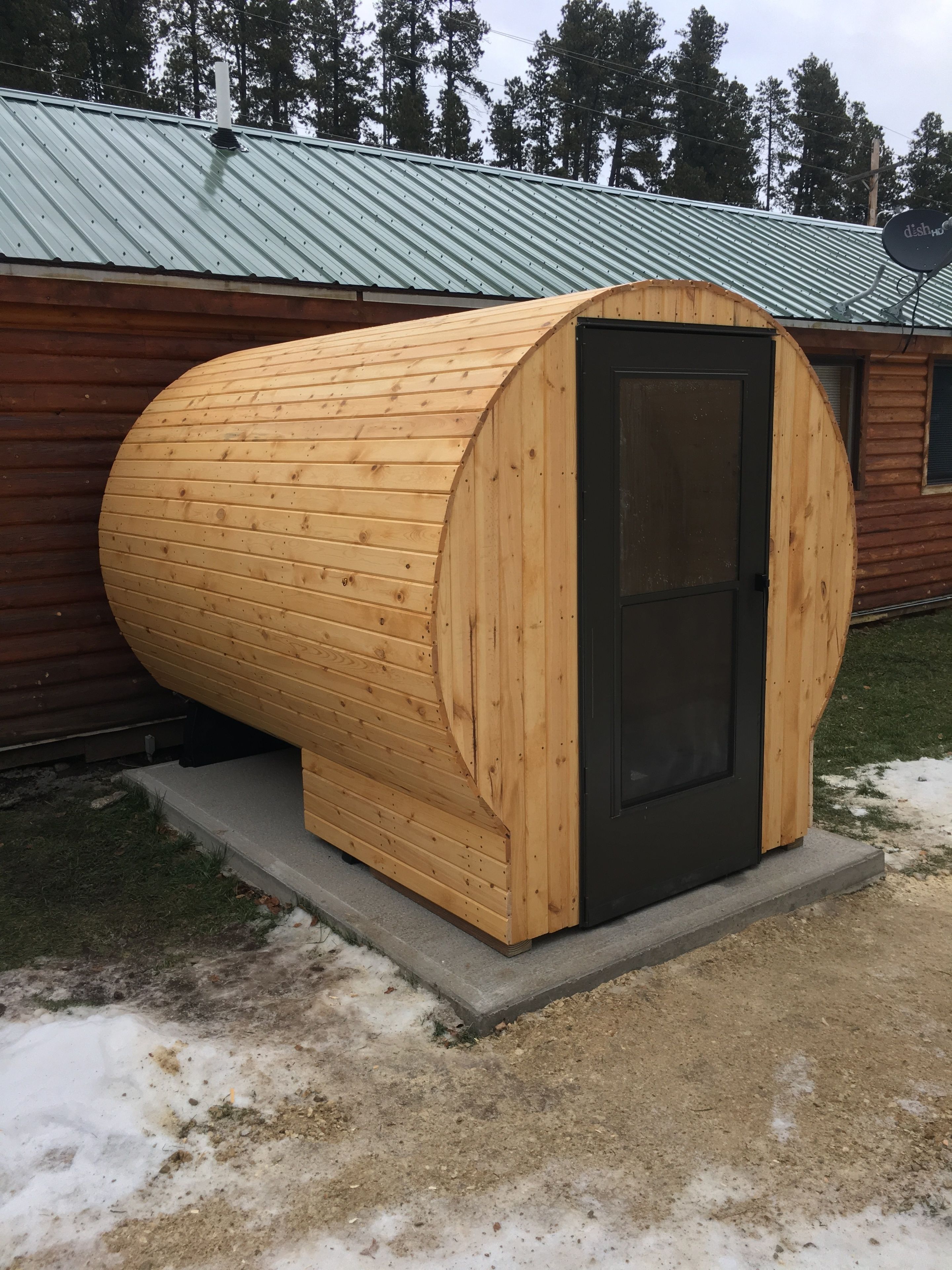A wooden sauna with a black door, situated outdoors next to a building with a metal roof.