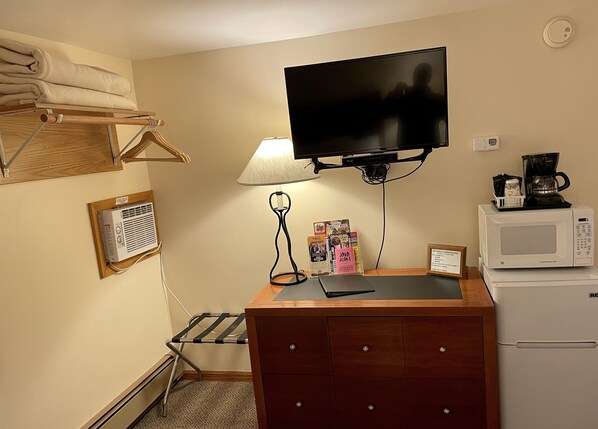 A compact hotel room with a flat-screen TV mounted above a wooden dresser, a microwave, a coffee maker, and a small table with magazines. - Whitetail Creek Resort (Lead)