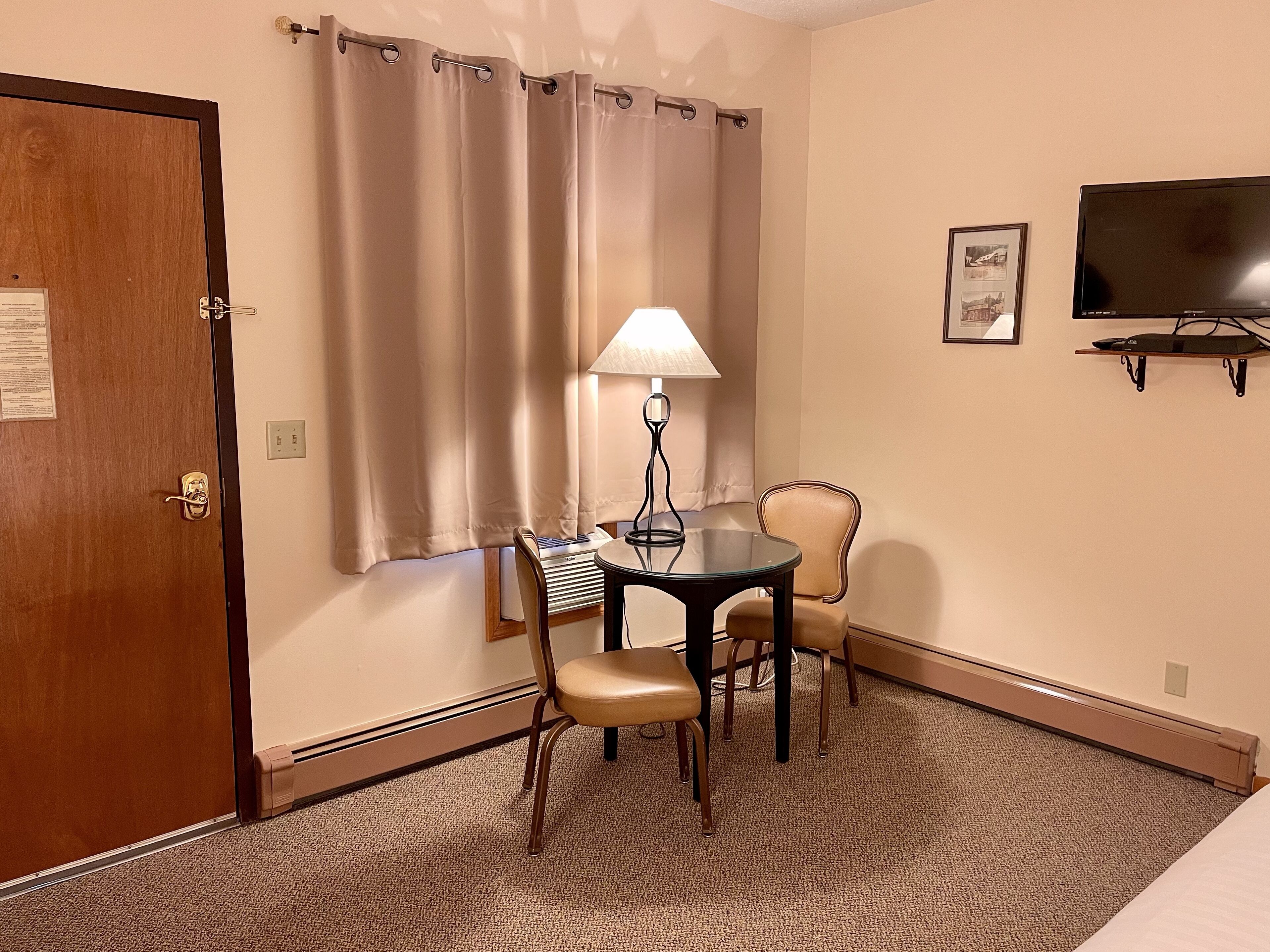 Une chambre d’hôtel avec une porte en bois, des rideaux beiges, une table avec une lampe, deux chaises, un téléviseur fixé au mur et un sol recouvert de moquette.
